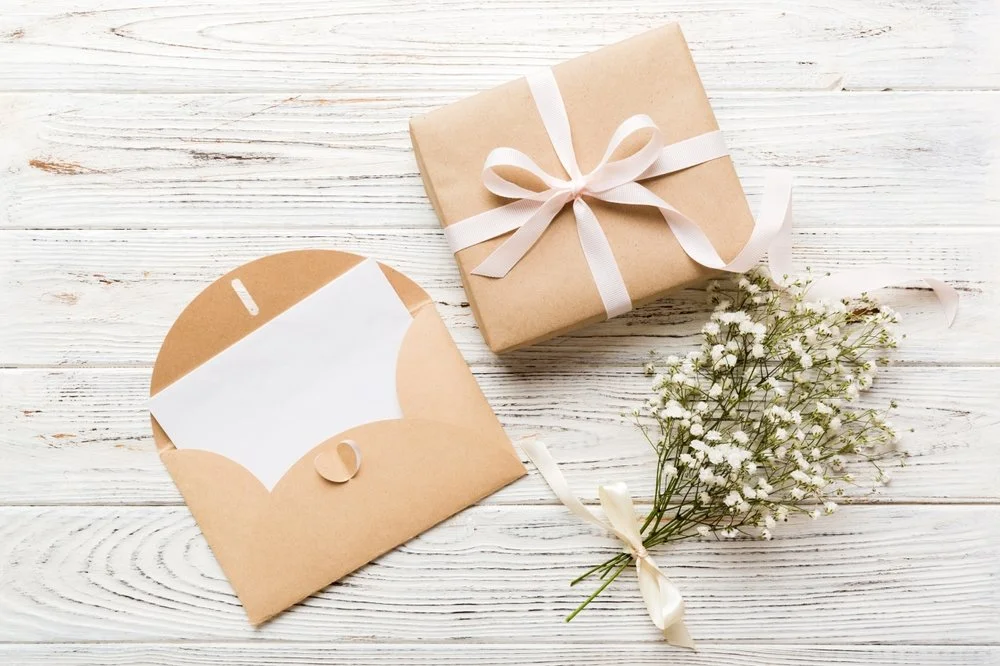 Un cadeau enveloppé dans du papier kraft avec un ruban blanc, une enveloppe en papier kraft, et une petite branche de fleurs blanches sur une table en bois blanc.