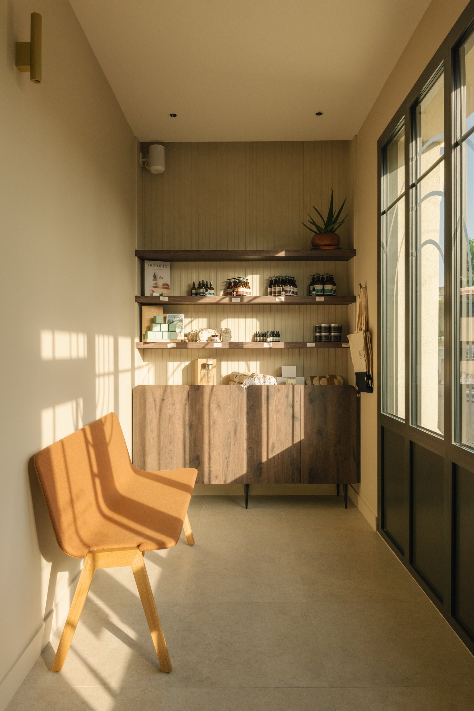Une salle avec une grande fenêtre laissant entrer la lumière naturelle, un fauteuil en bois beige, une étagère en bois avec des produits de soins et une plante dans un pot en terre cuite placée sur une étagère supérieure. Accueil Spa Beroa.