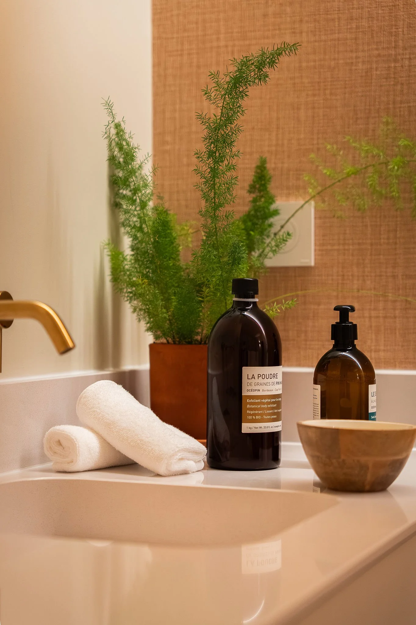 Un cuarto de baño con una planta verde en una maceta, dos botellas de productos de aseo, toallas blancas enrolladas, una bañera blanca, un lavabo con un grifo, un cuenco de madera y una pared con papel pintado marrón.