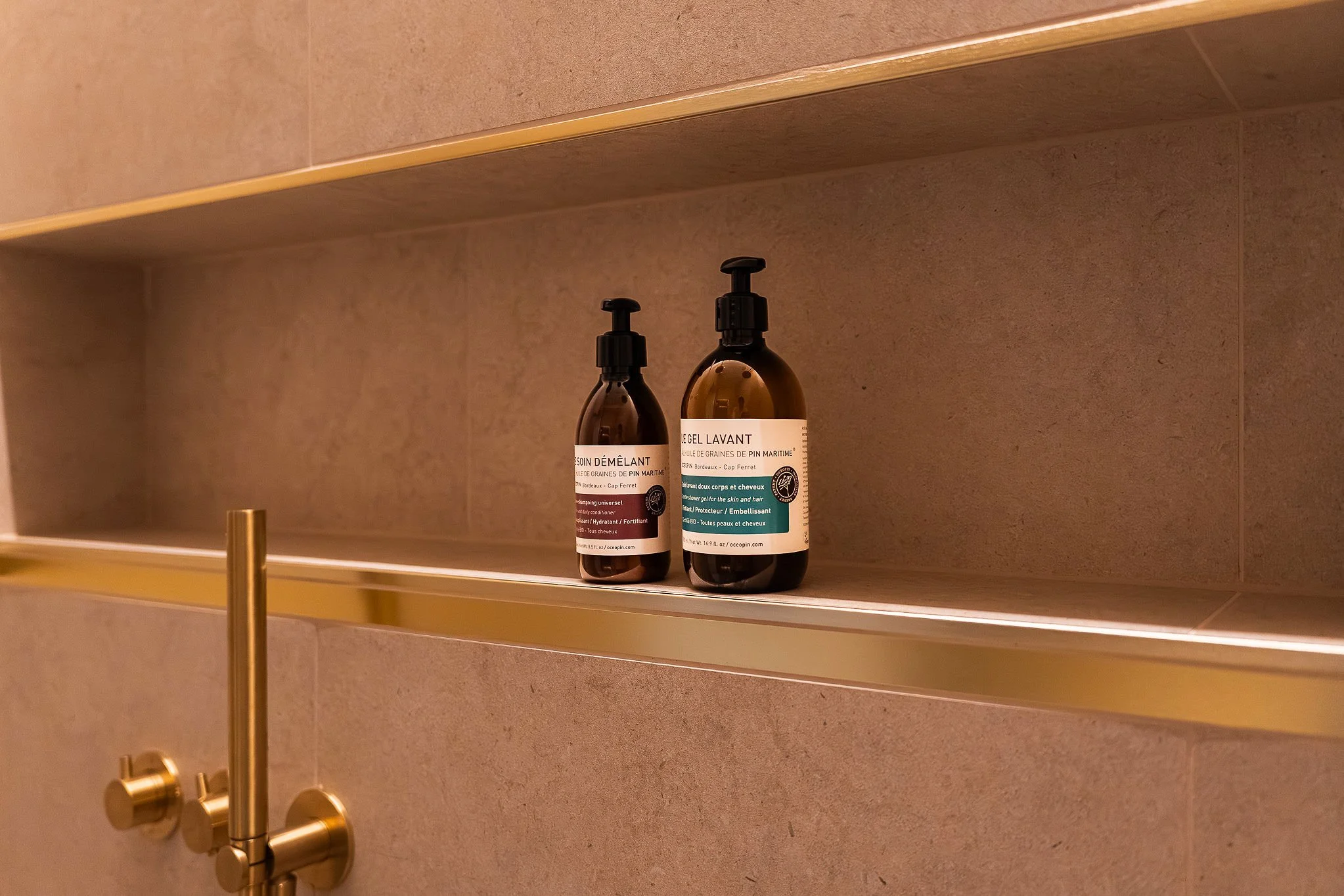 Dos frascos de productos para el cuidado de la piel y el cabello en una estantería de un cuarto de baño, con una pared de azulejos beige al fondo.
