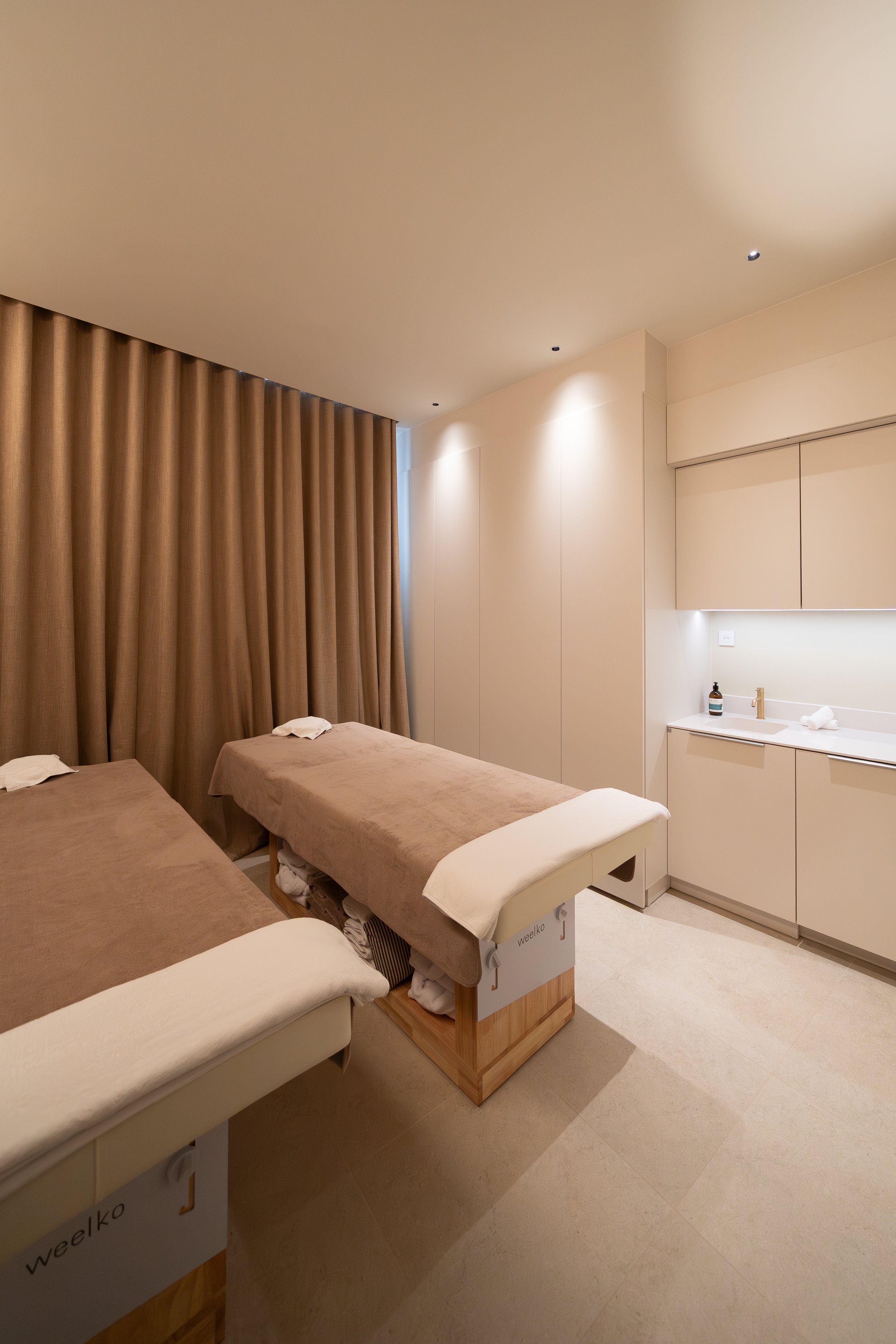 Une salle de massage ou de soin avec des tables recouvertes de draps beiges, un mur beige clair, des rideaux marron, une vasque avec un savon et une lotion repose sur un comptoir blanc, dans un environnement calme et épuré. Salle Duo Cabine Spa Beroa