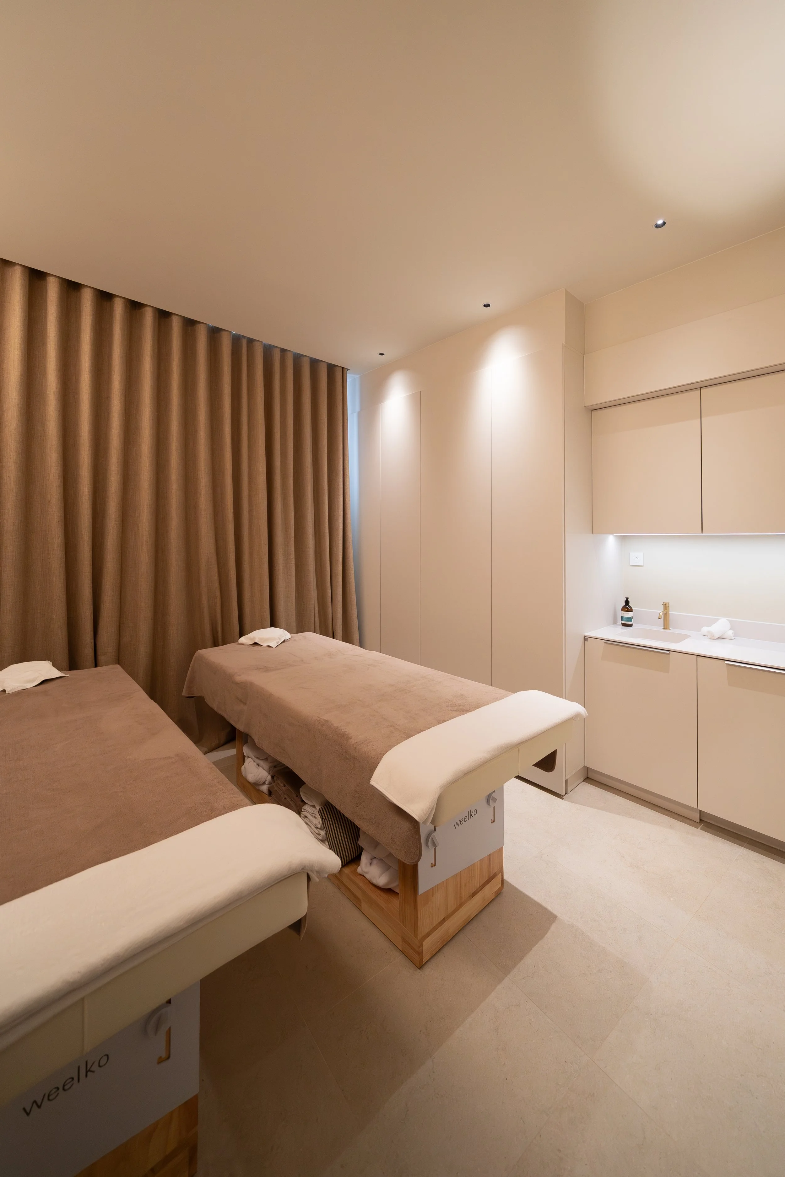 Salle de massage avec trois tables de massage en beige, grande pièce lumineuse avec murs crème, rideaux beiges, éclairage encastré et un coin lavabo avec savon, serviettes et vasque.