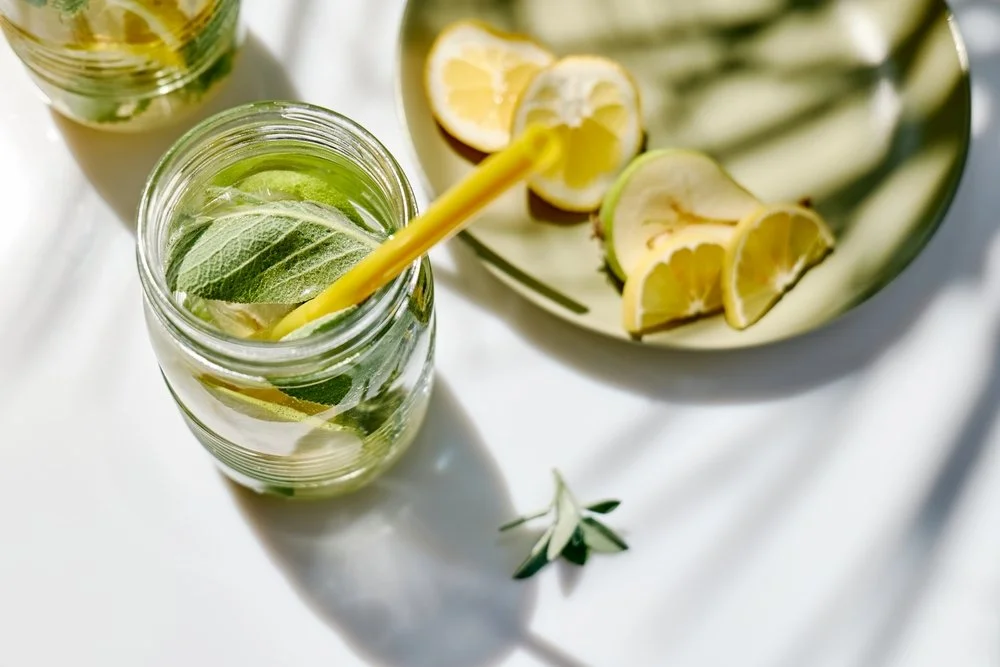 Un verre mason rempli d'eau, de feuilles de menthe, de citron et de concombre, avec une paille jaune, sur une surface blanche, à côté d'une assiette avec des quartiers de citron et de citron vert.