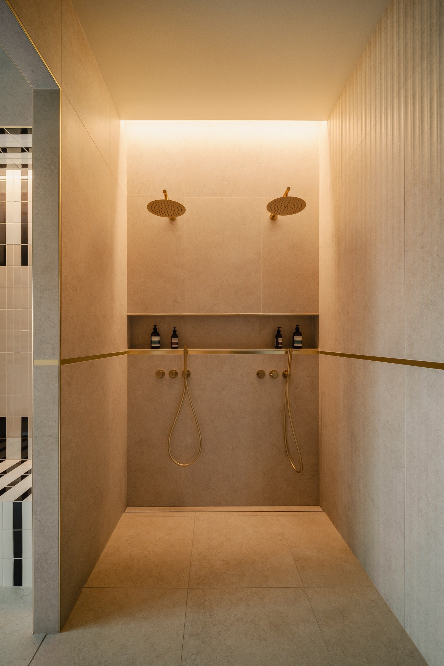 Deux douches avec pomme de douche ronde en cuivre, chaque douche ayant un robinet et un tuyau en cuivre suspendu, dans une cabine de douche en pierre beige avec un éclairage doux en haut.  Spa Beroa.
