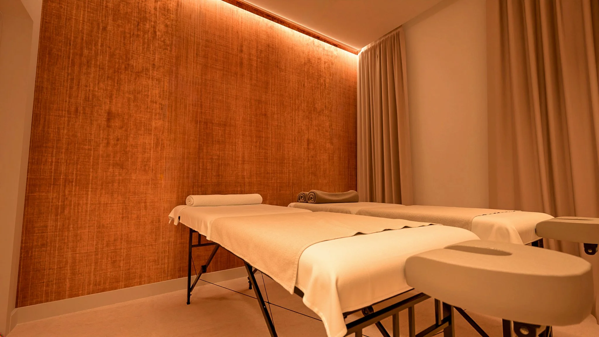 Salles de massage avec deux tables à massage recouvertes de draps blancs et plusieurs serviettes roulées, dans une pièce chaleureuse avec un mur orange et des rideaux beige.