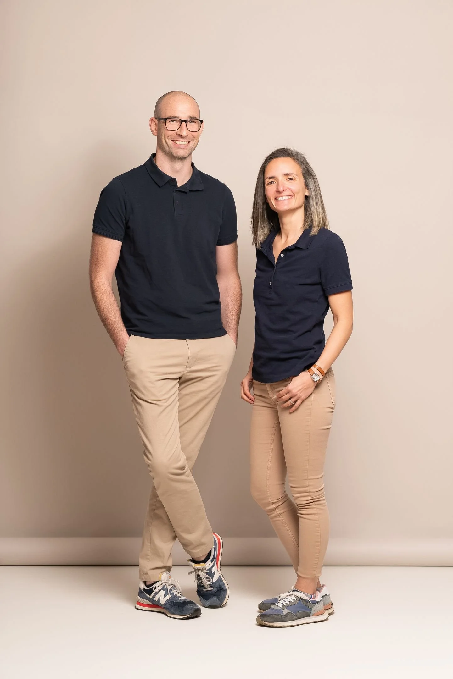 Deux personnes posant côte à côte contre un mur beige, portant des vêtements décontractés et des chaussures de sport, souriant à la caméra.