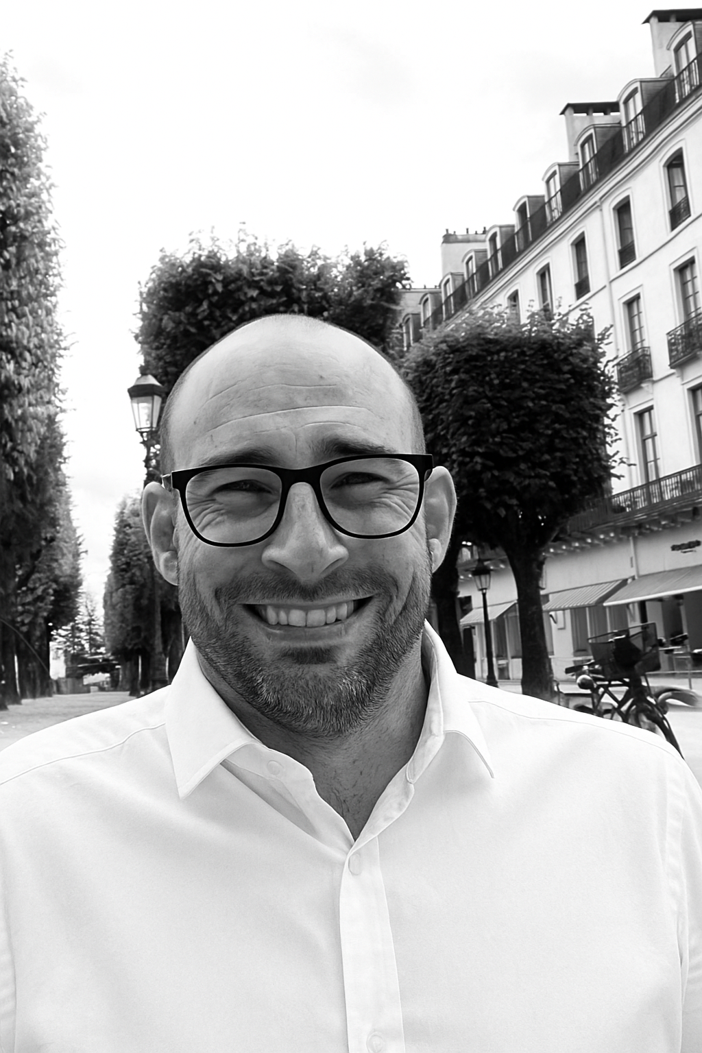 Un homme souriant portant des lunettes noires et une chemise blanche, debout dans une rue bordée d'arbres et d'immeubles résidentiels, en noir et blanc.