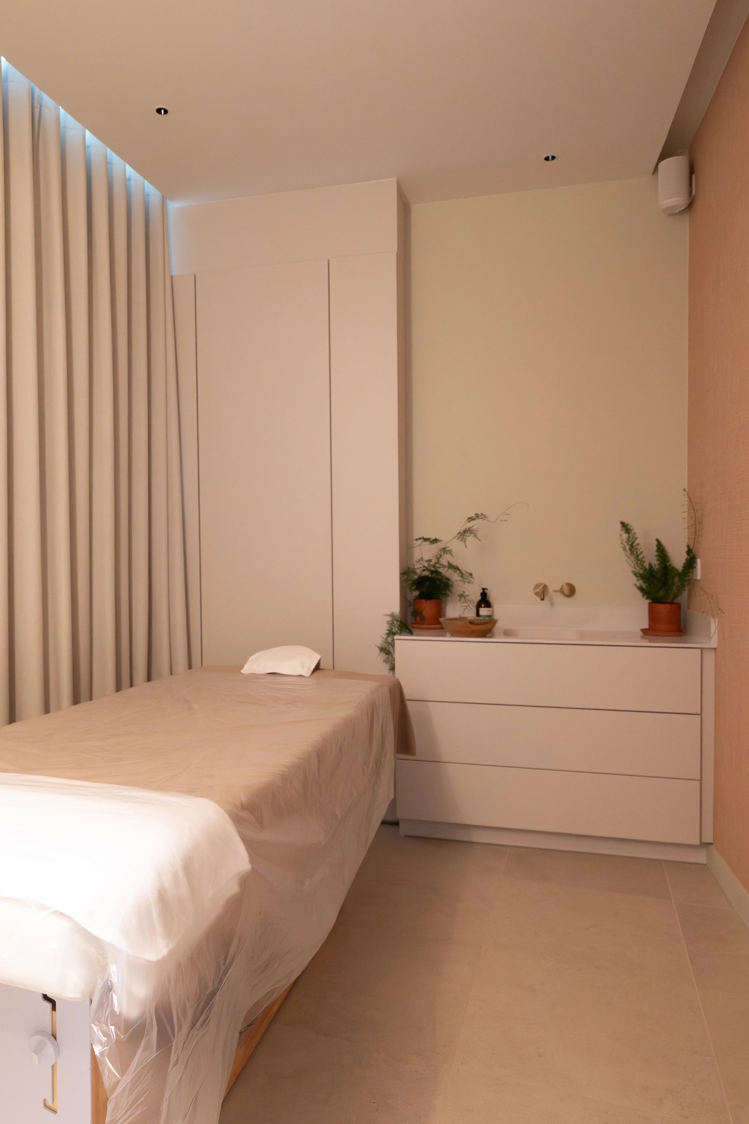 Chambre minimaliste avec lit, drap blanc, oreiller, meuble blanc avec plantes vertes et décorations naturelles, rideaux beige, murs blanc et beige, sol en tuiles beige clair.