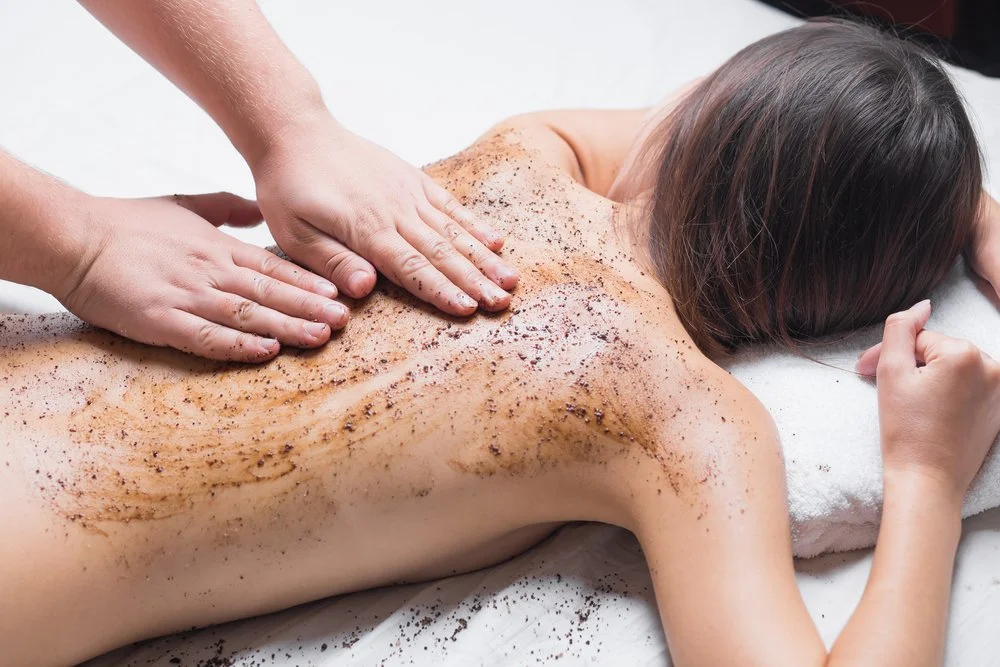 Une personne reçoit un massage dans un spa, avec des exfoliants ou des gommages appliqués sur le dos. La personne est allongée sur une table de massage avec une serviette blanche.