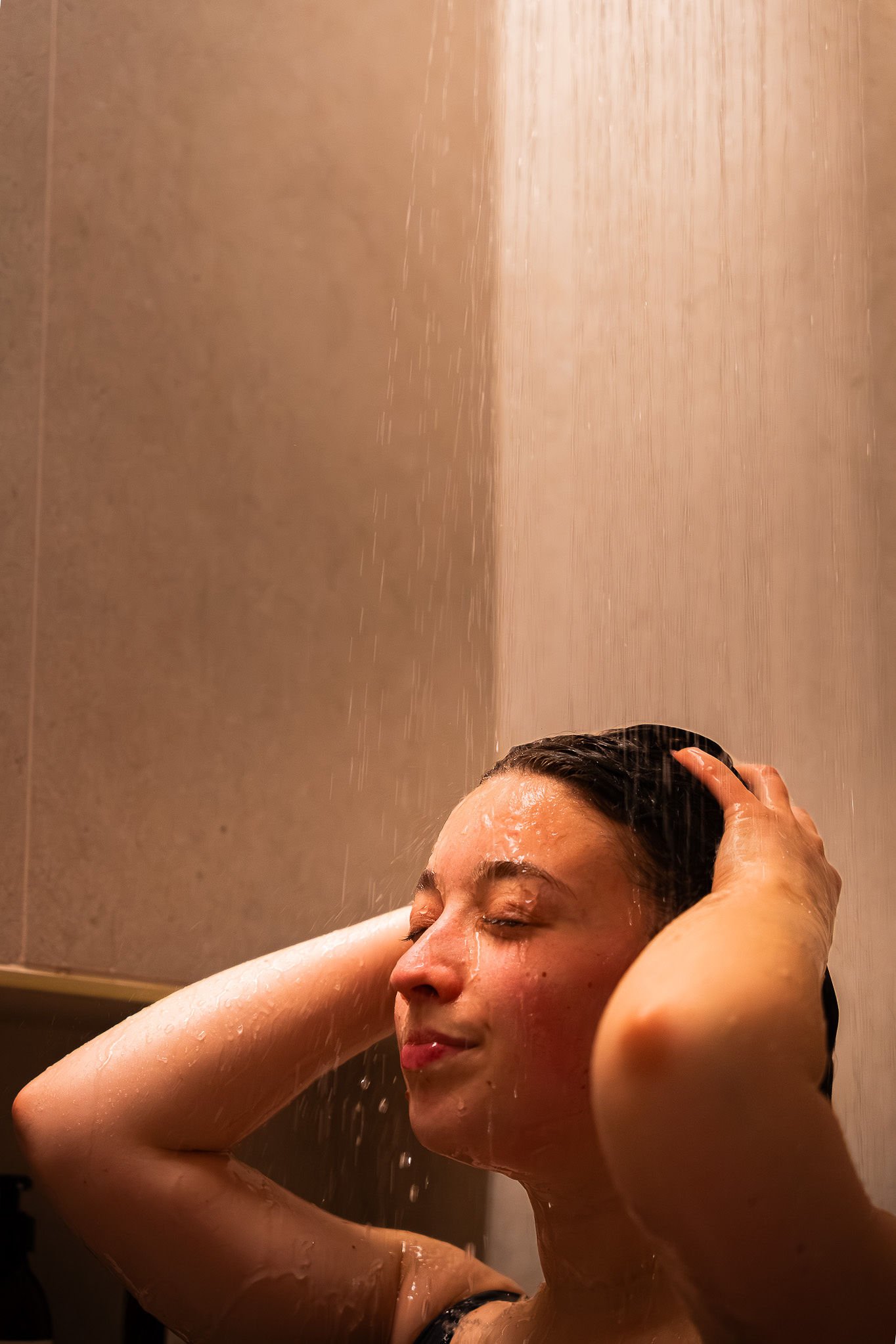Une femme prend une douche, elle a les yeux fermés, les mains dans ses cheveux, et de l'eau coule sur son visage et ses épaules au Spa Beroa.