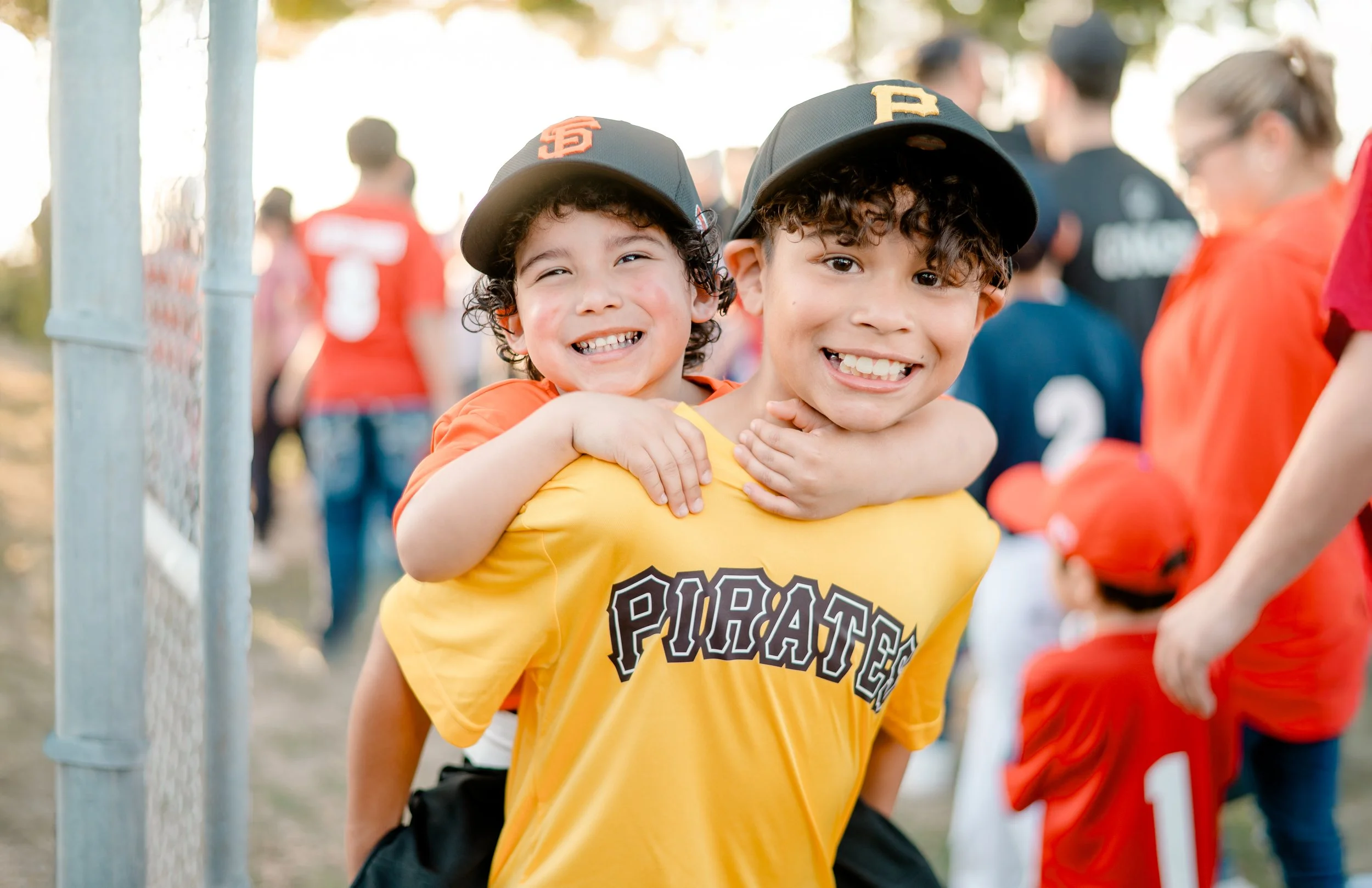 Two young boys at a baseball game, one giving the other a piggyback ride, both smiling, wearing Pittsburgh Pirates hats and jerseys, surrounded by other game attendees.