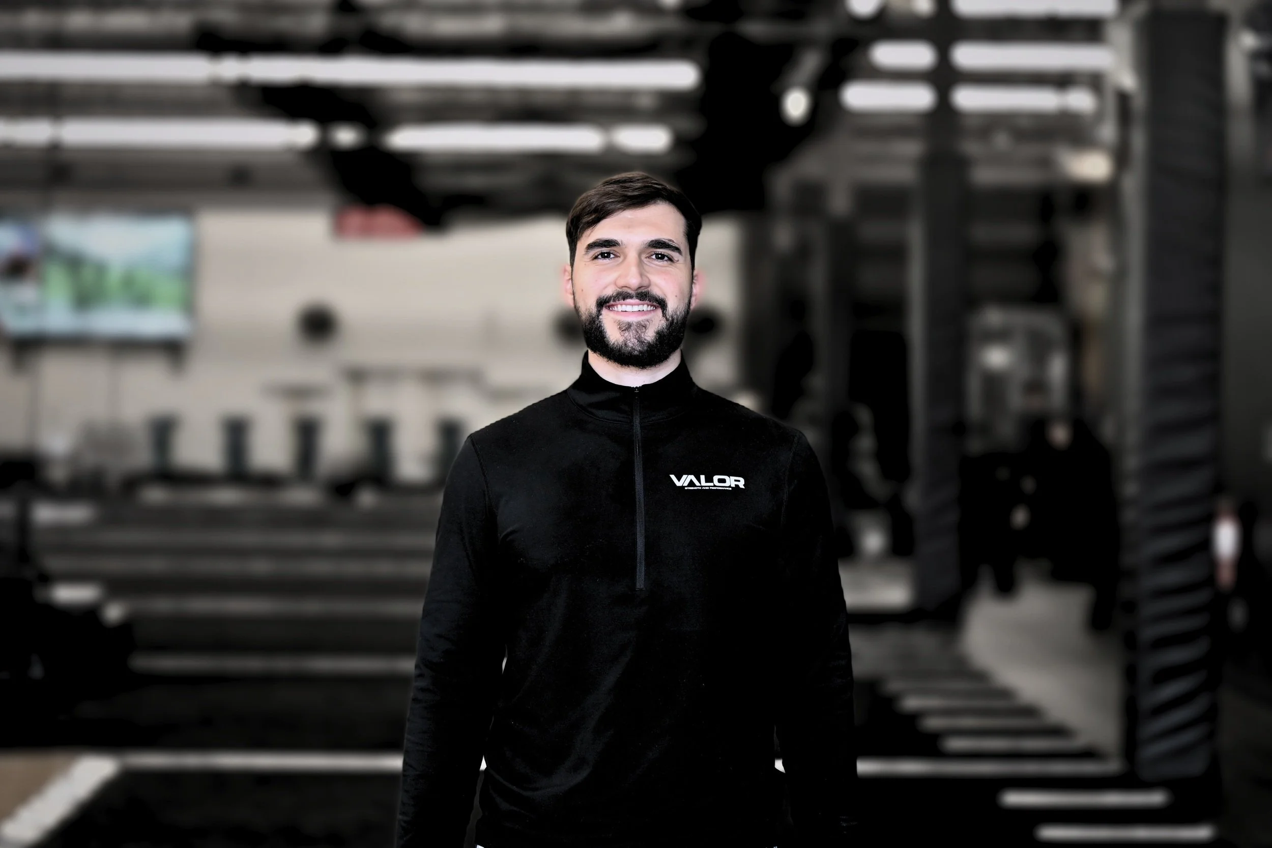 A smiling man with dark hair and beard wearing a black athletic jacket with 'VALOR' logo, standing in a gym with fitness equipment in the background.