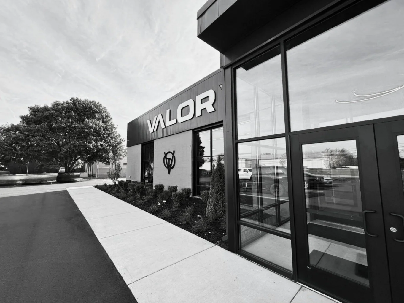 Exterior of a building with the sign 'VALOR' on it, featuring glass doors and windows, with a parking lot, trees, and a clear sky in the background.