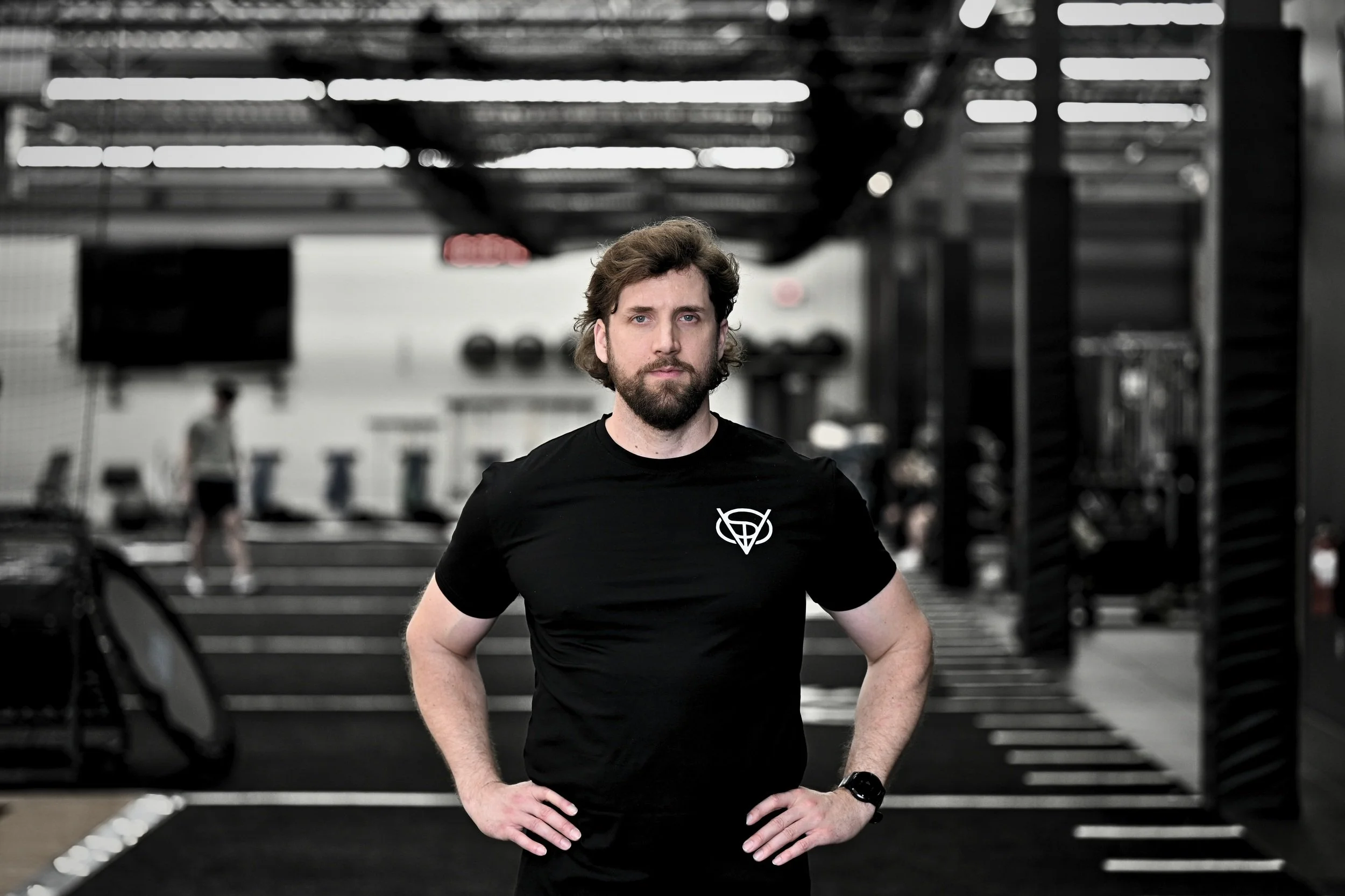 A man with brown hair and a beard in a black t-shirt standing with hands on hips inside a gym.