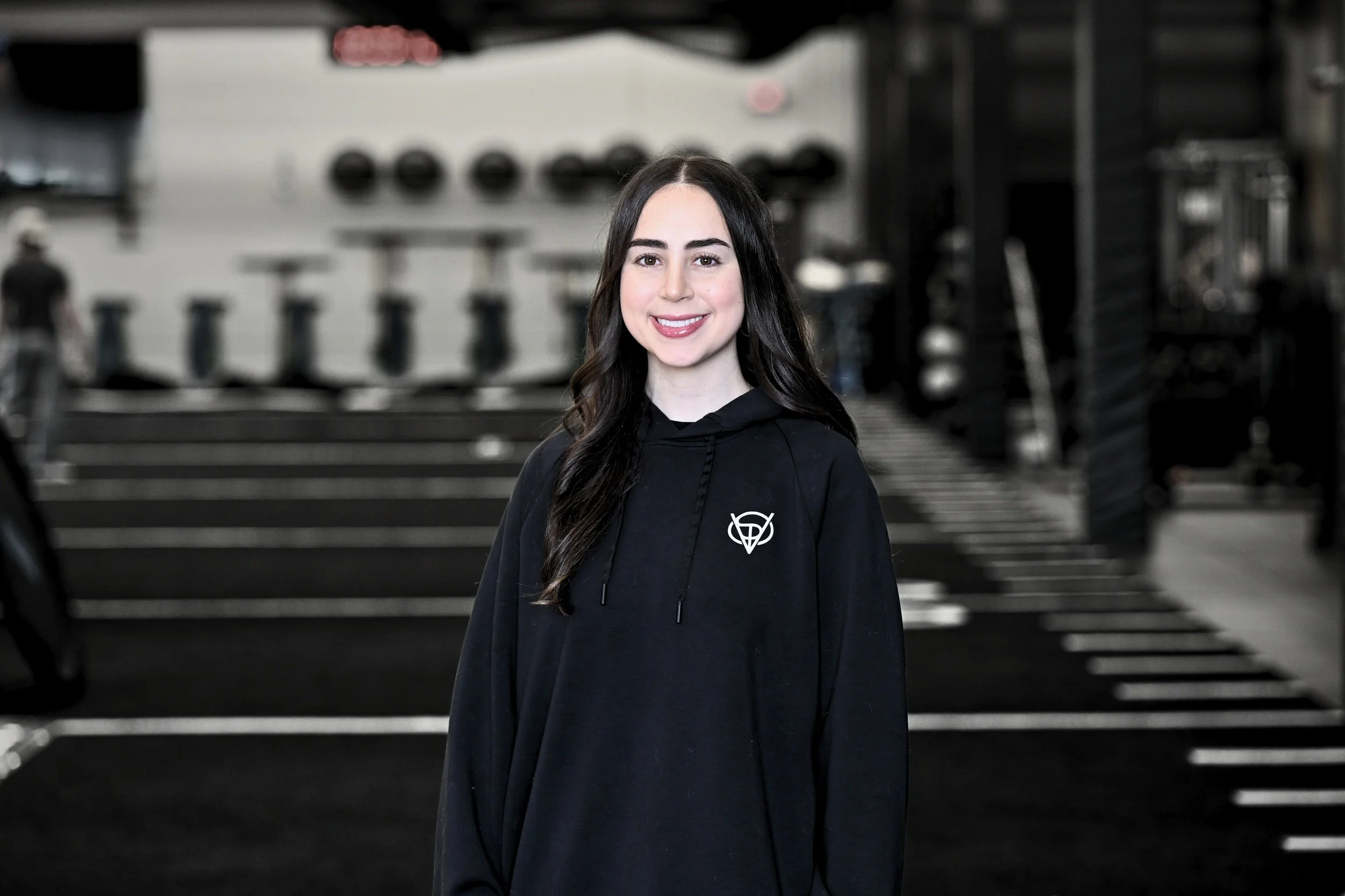 A young woman with long dark hair, smiling, wearing a black hoodie with a white logo, standing in a gym.