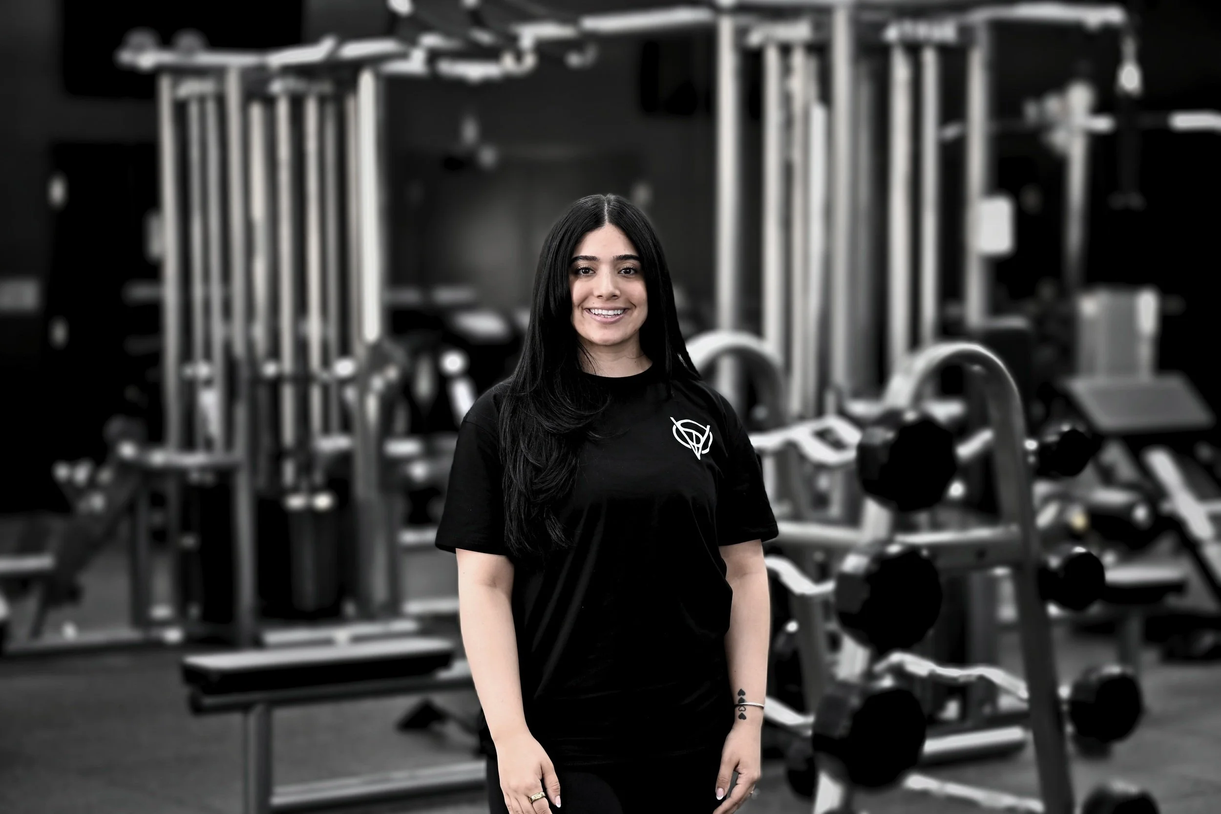 A smiling woman with long black hair standing in a gym surrounded by workout equipment.