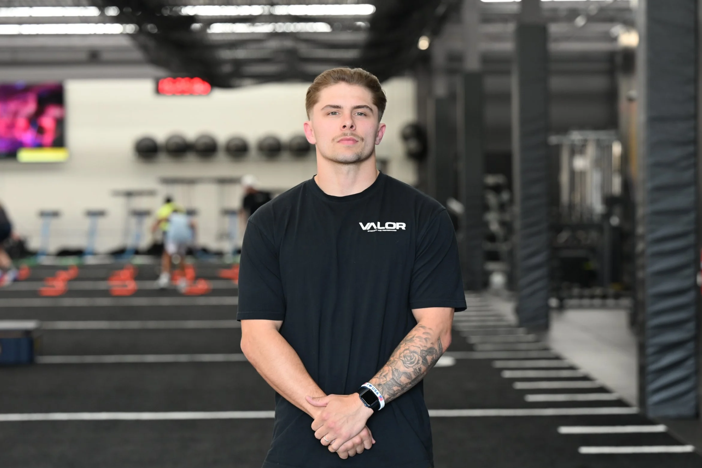 Young man standing in a gym, wearing a black T-shirt with 'VALOR' logo, with gym equipment and other people working out in the background.