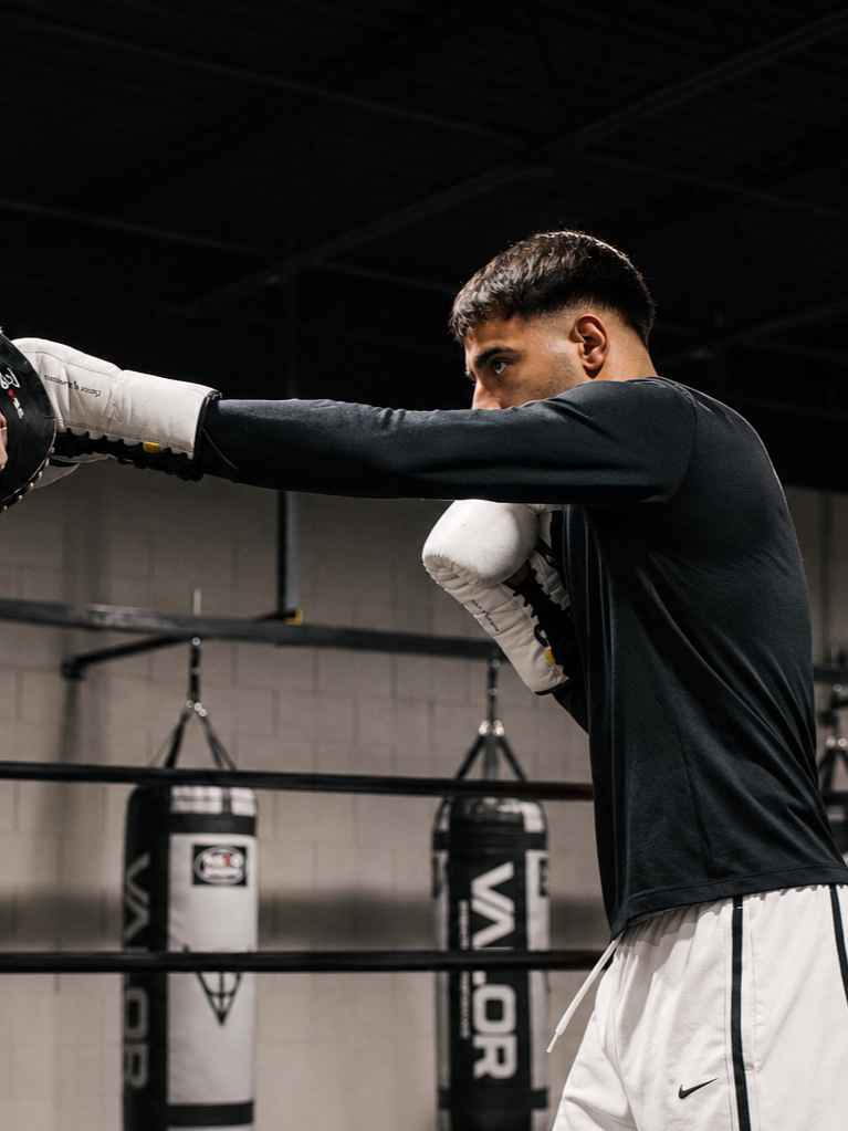 Young male boxer in black shirt and white shorts training in boxing gym, practicing punches with gloves on, boxing bags in background.