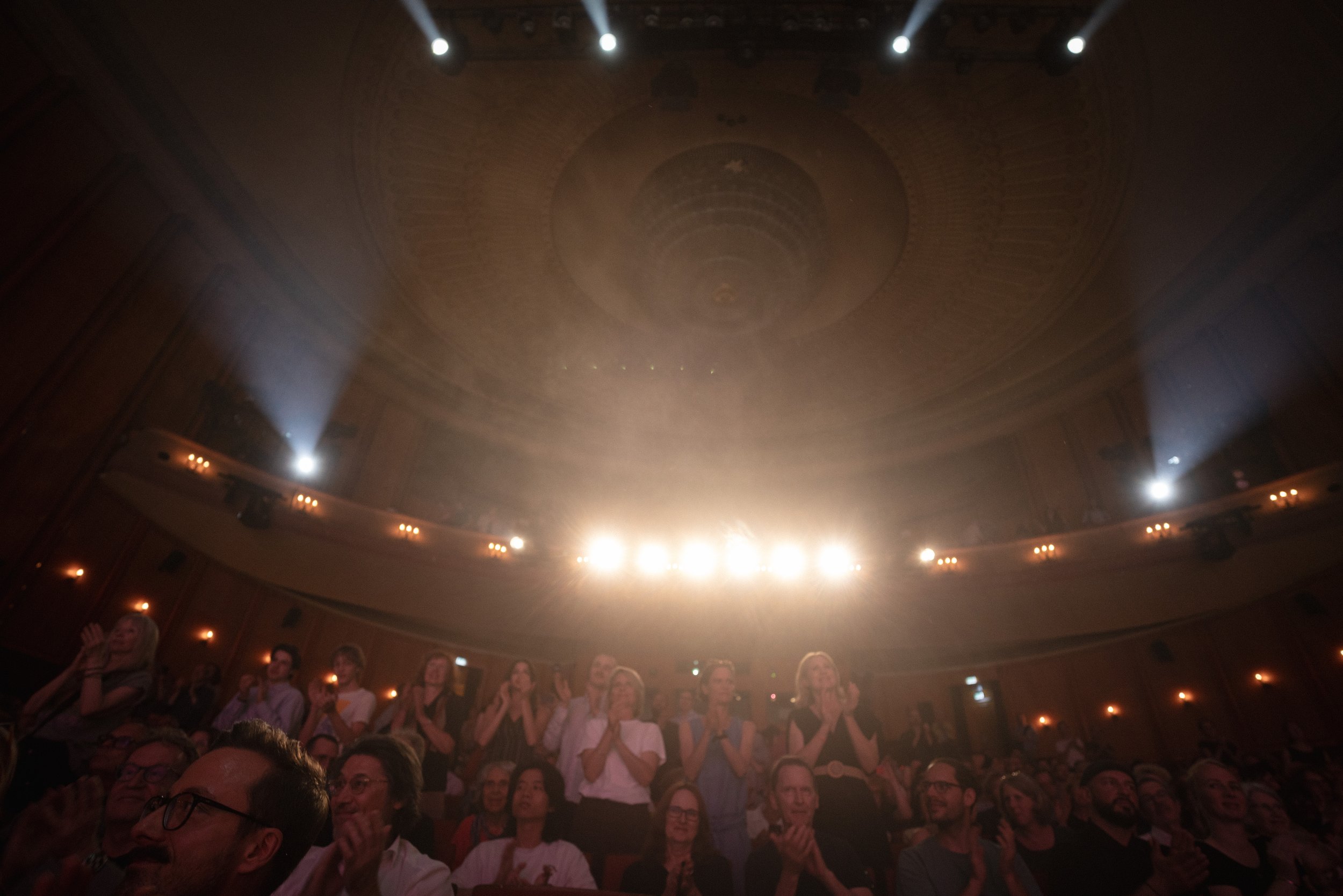 Menschen sitzen und stehen in einem Theater oder einer Konzerthalle, während auf der Bühne Scheinwerfer leuchten.