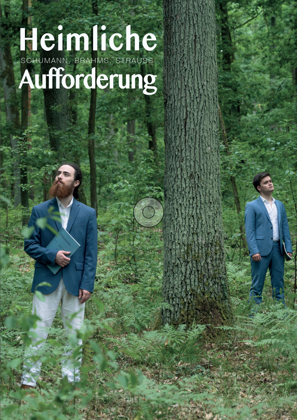 Zwei Männer in Anzügen stehen in einem grünen Wald, einer hält ein Buch, während sie nach oben schauen.