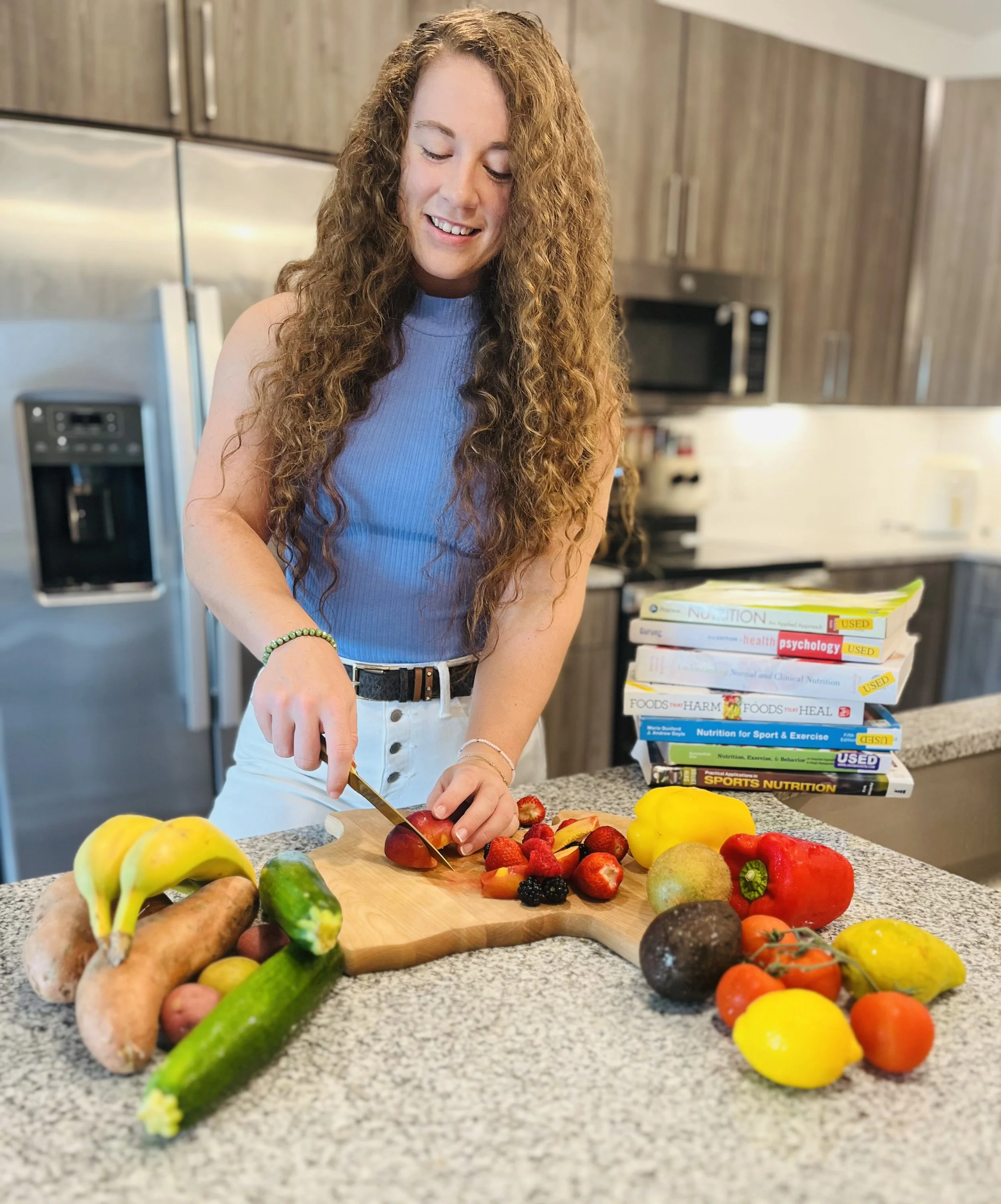 A woman with long curly red hair slicing a strawberry in a kitchen with a variety of fresh fruits on the counter and a stack of books nearby.