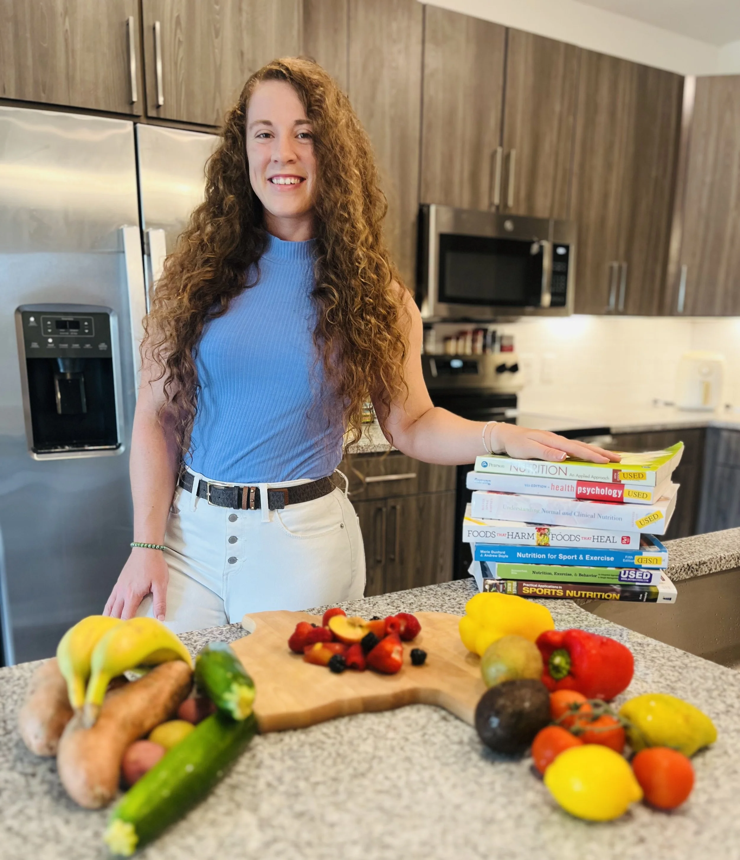 A woman with long curly hair wearing a blue sleeveless top and white pants, standing in a kitchen, smiling, with a stack of health and nutrition books on the counter beside her, surrounded by fresh fruits and vegetables.