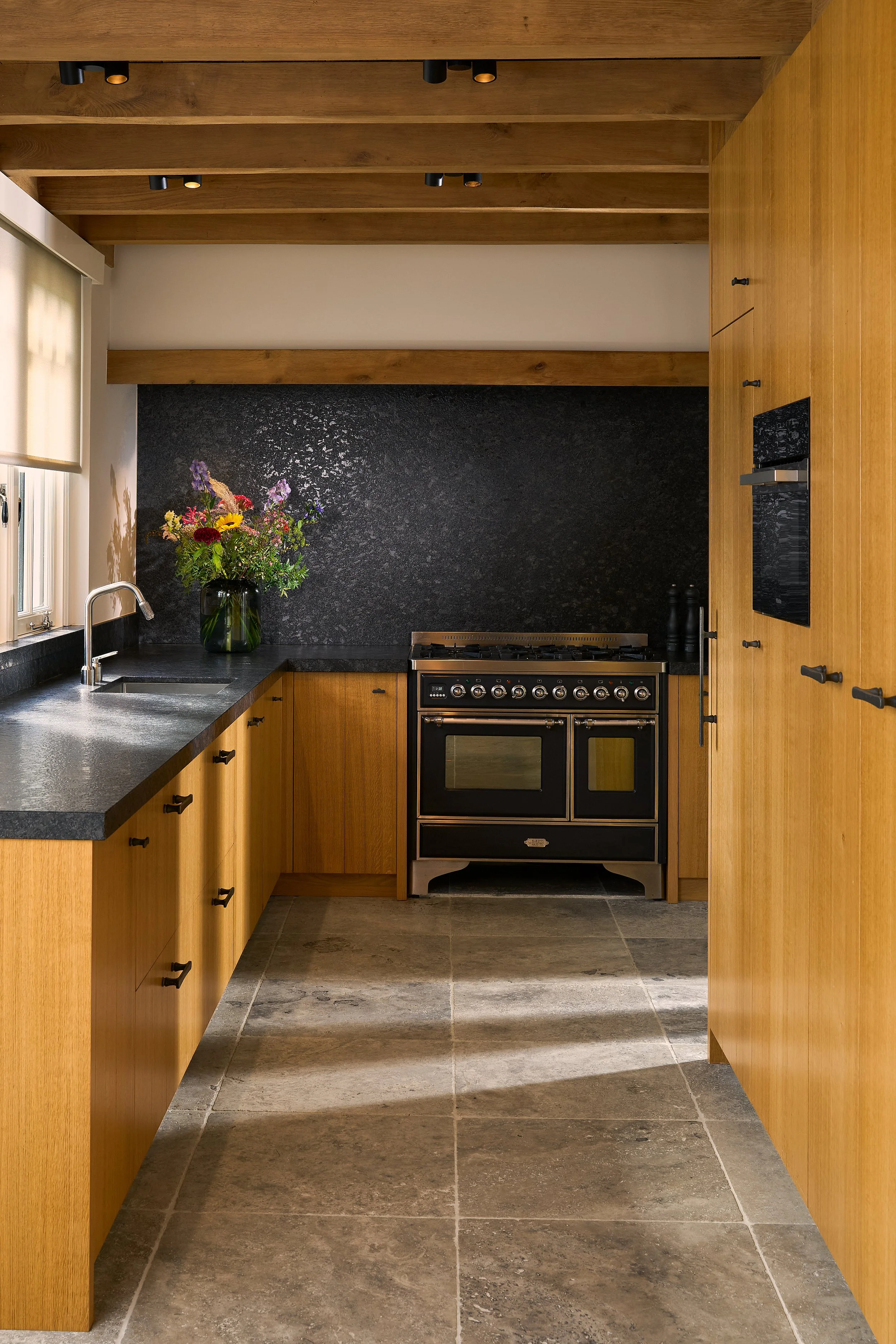 A modern kitchen with wooden cabinets, a black countertop, a black stove, and a black backsplash. There is a window with a beige blinds and a vase with flowers on the countertop. The ceiling has exposed wooden beams with black recessed lighting, and