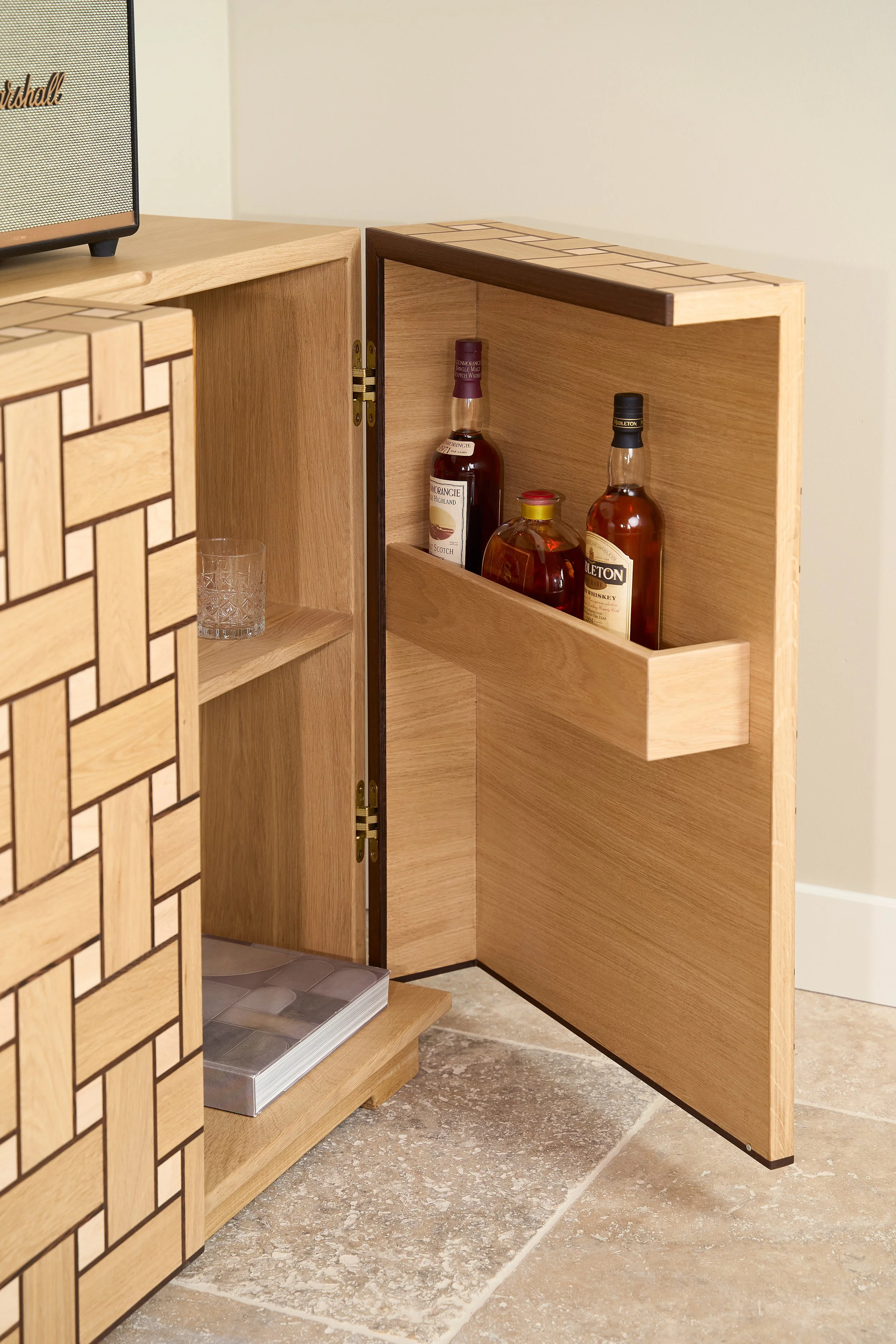 Open wooden cabinet with bottles of liquor inside, two on the top shelf and one on the middle shelf, with glassware and a hardbound book on the bottom shelf.