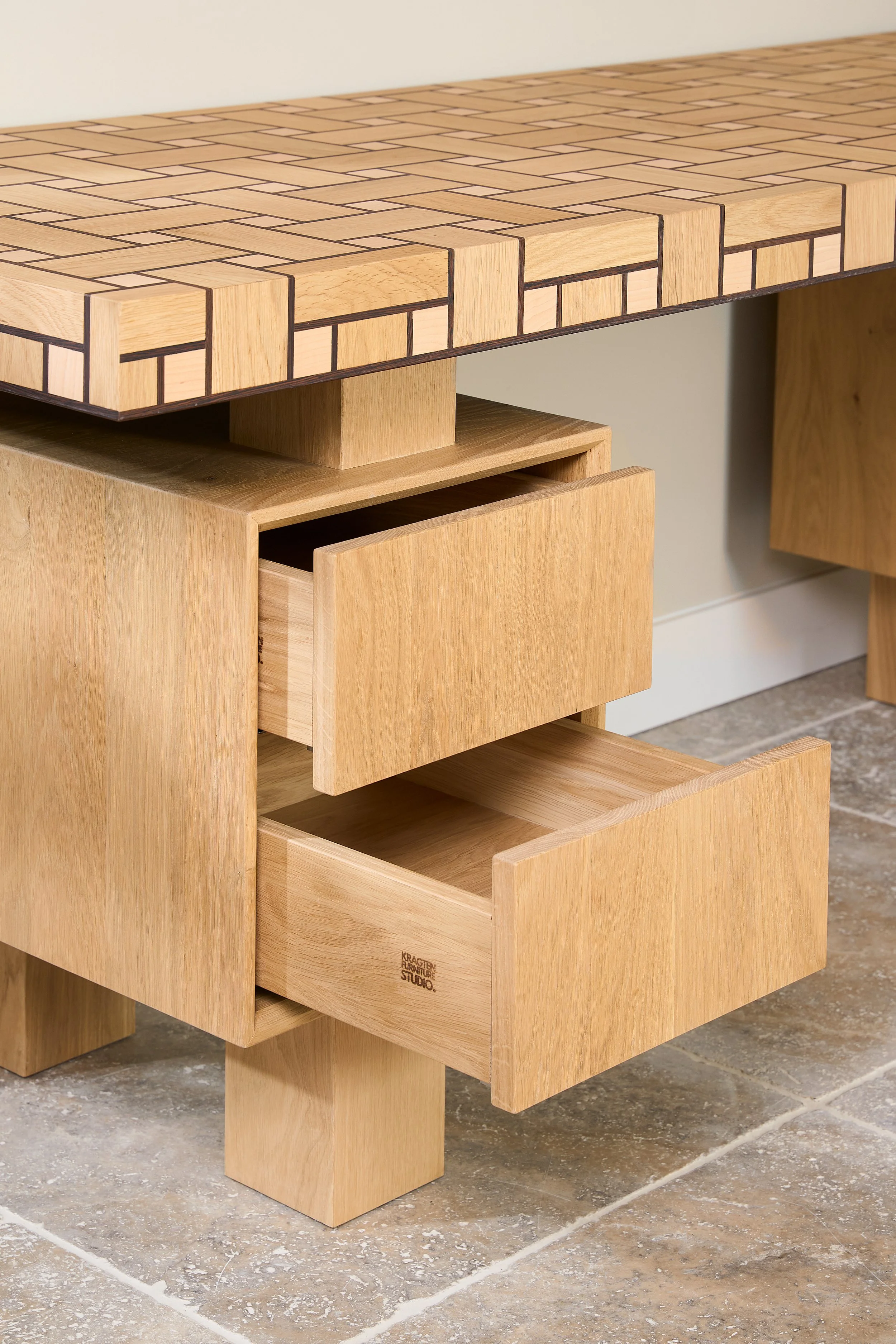 Wooden desk with a patterned top and two open drawers, situated on a tiled floor.