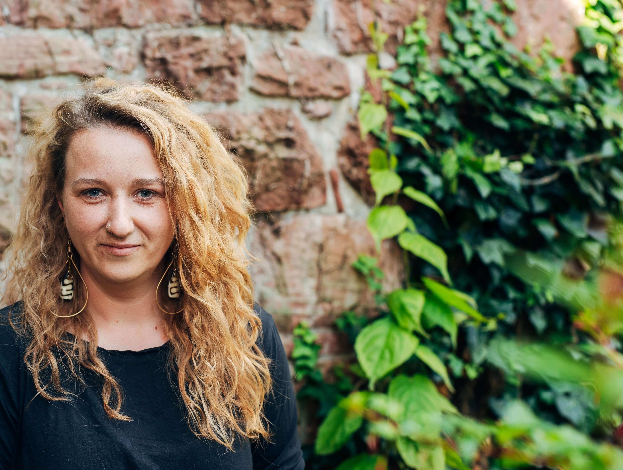 Eine junge Frau mit lockigem, blondem Haar, schwarzem Oberteil und großen Ohrringen steht vor einer Steingartenmauer und grünen Pflanzen.