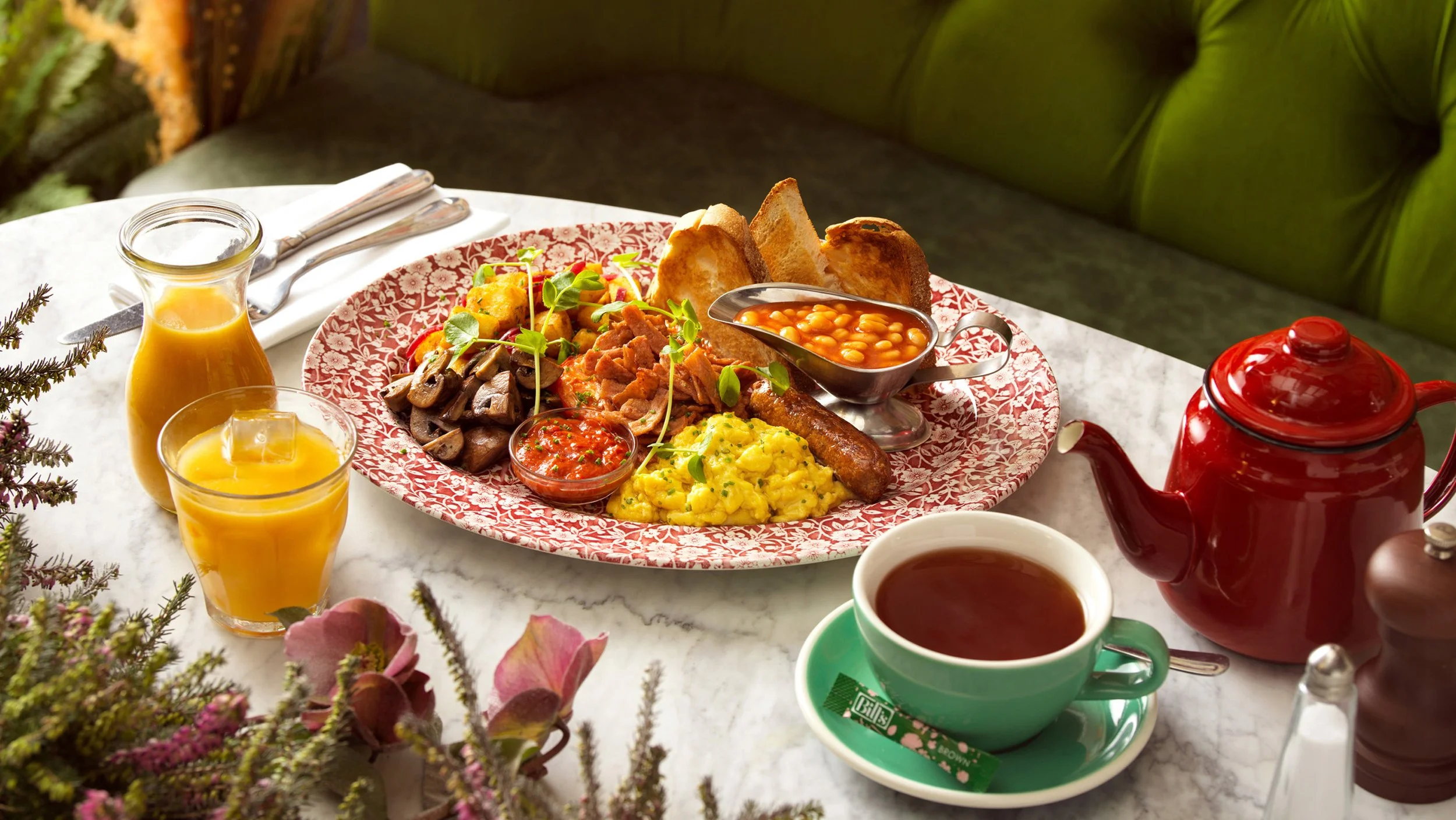 Traditional English breakfast with baked beans, scrambled eggs, sausage, mushrooms, tomatoes, toast, and accompaniments including orange juice, tea, and a teapot, served on a marble table with flowers.