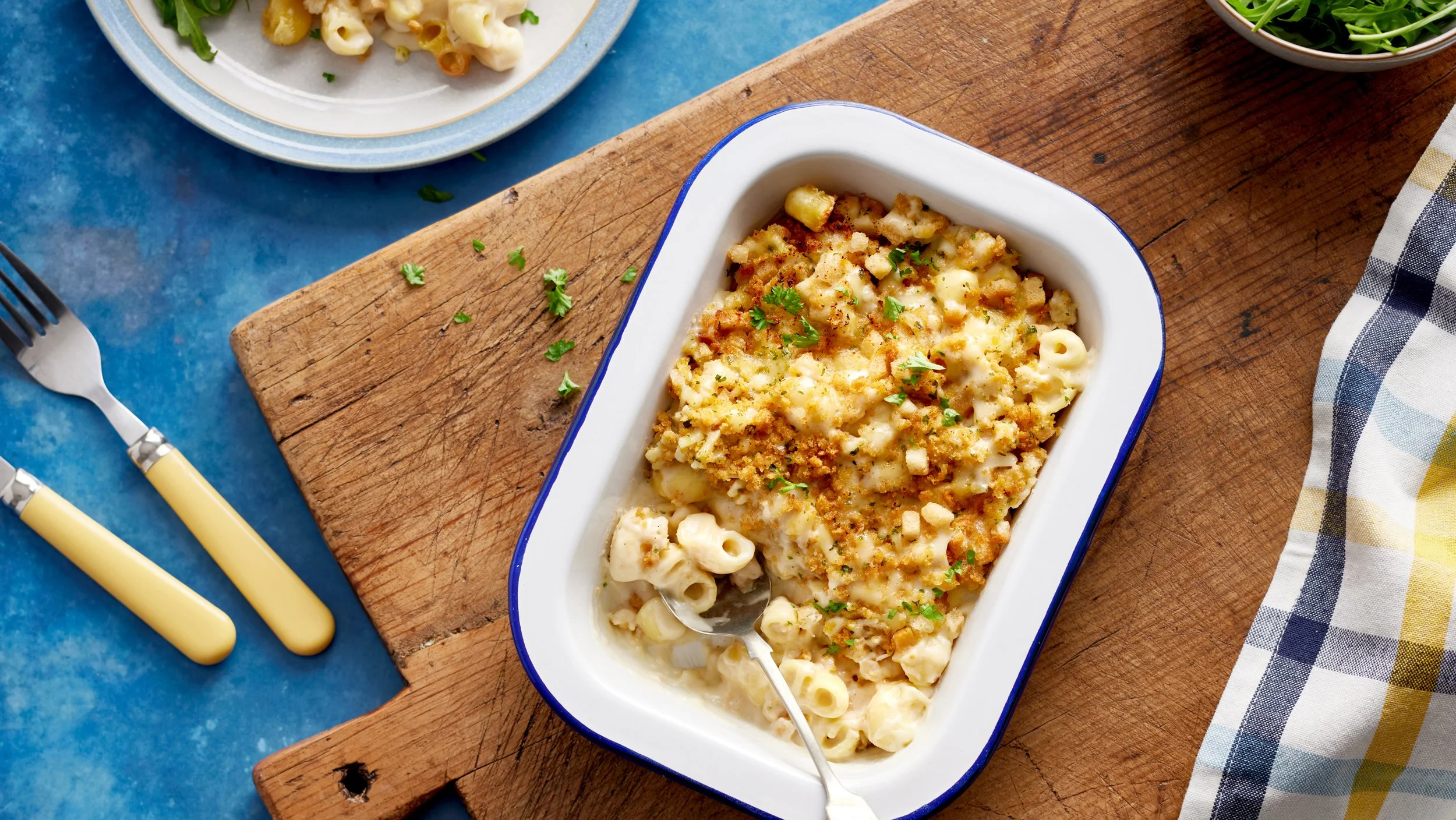 Baked macaroni and cheese topped with breadcrumbs, garnished with chopped herbs in a white and blue dish on a wooden cutting board, with a fork in the dish.