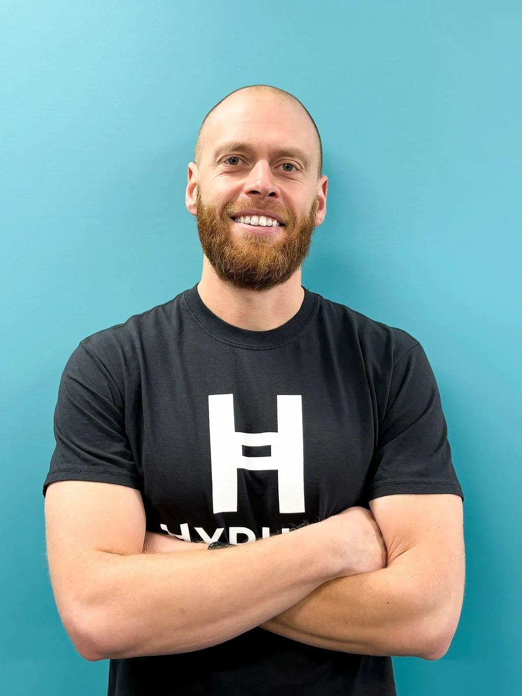 A man with a beard and a shaved head speaking passionately at what appears to be a gym or fitness class, wearing a dark hoodie with a small geometric logo.