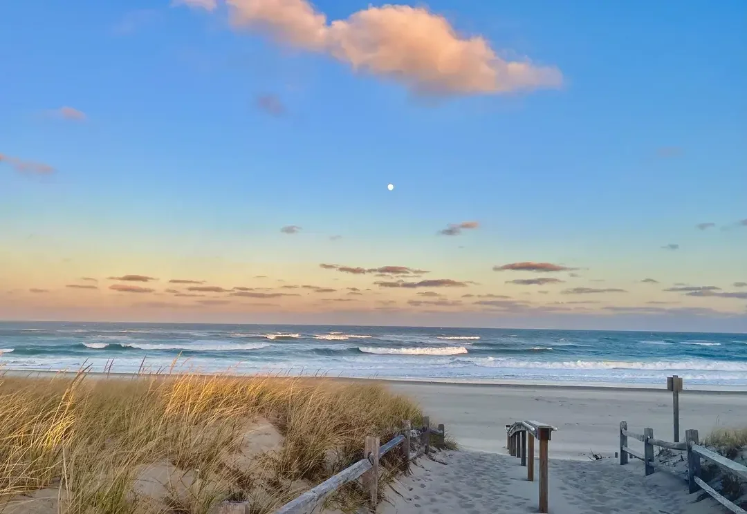 A sandy beach with grassy dunes and a wooden fence leading to the ocean, under a partly cloudy sky with the moon visible.