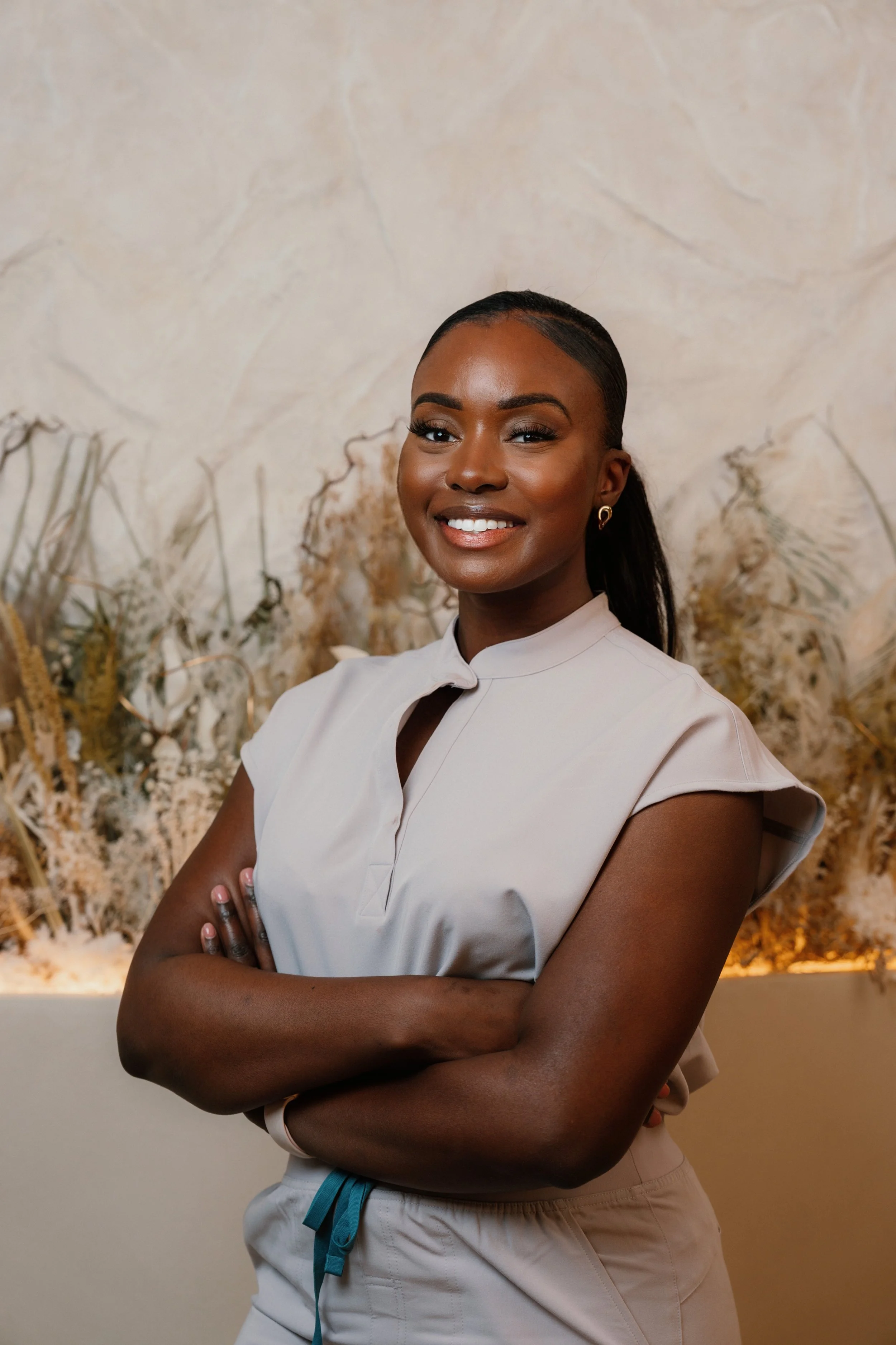 A female doctor with dark hair, smiling, wearing beige scrubs with short sleeves, standing with her arms crossed in front of a beige background with dried plants.