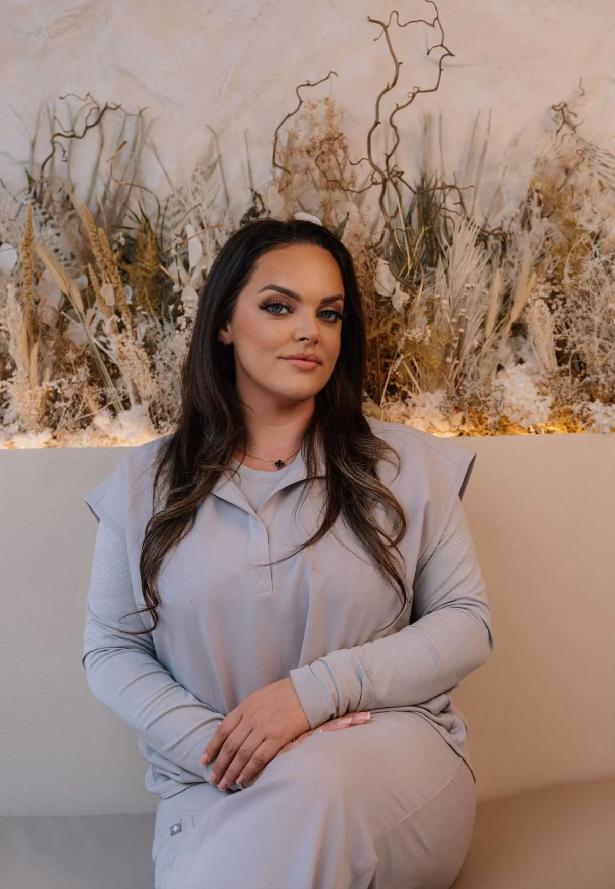 A woman with long brown hair, wearing beige scrubs, sitting against a neutral-colored background with dried decorative plants and branches.