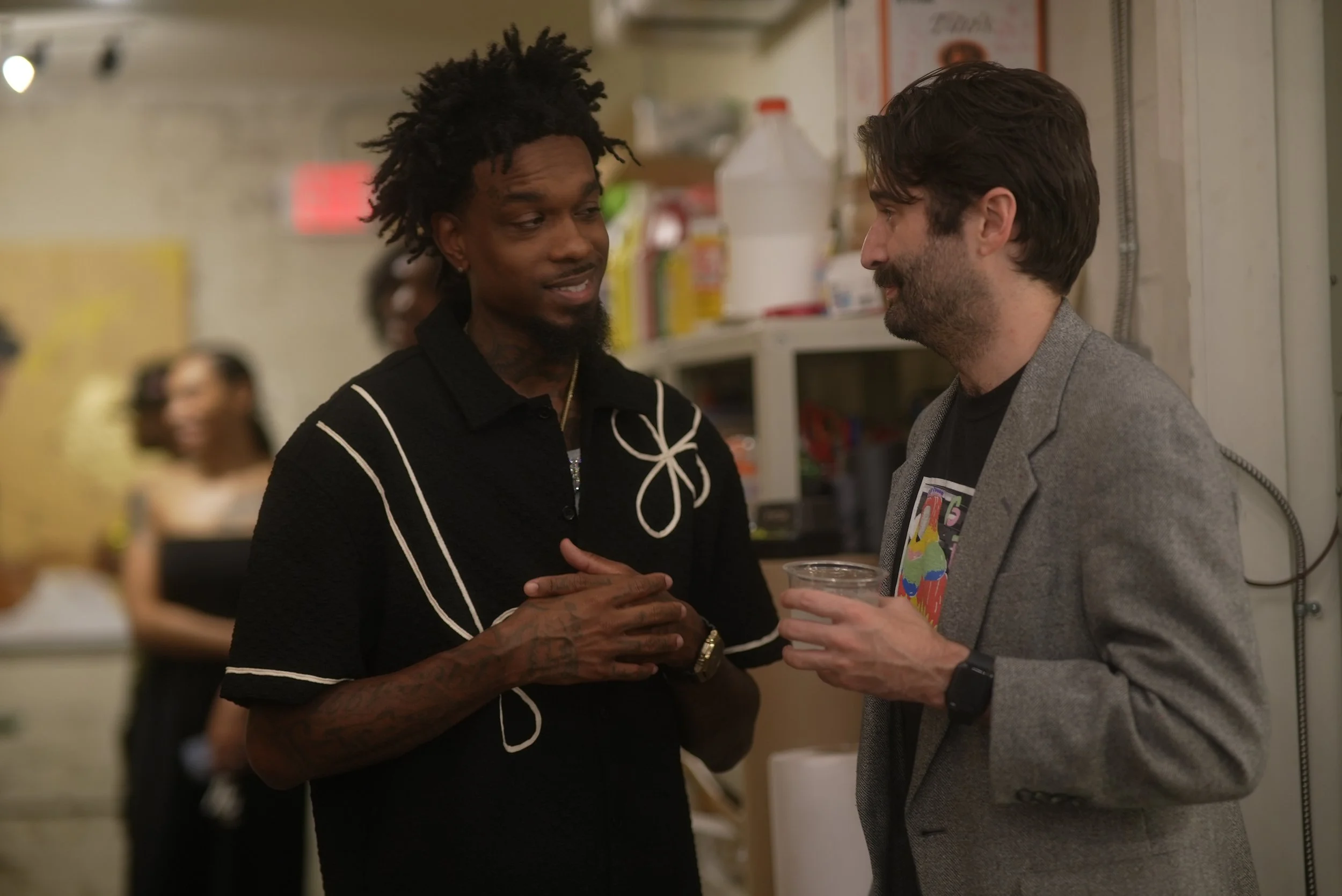 Two men engaged in conversation at a social gathering, one holding a drink. The background includes a woman smiling and shelves with various items.