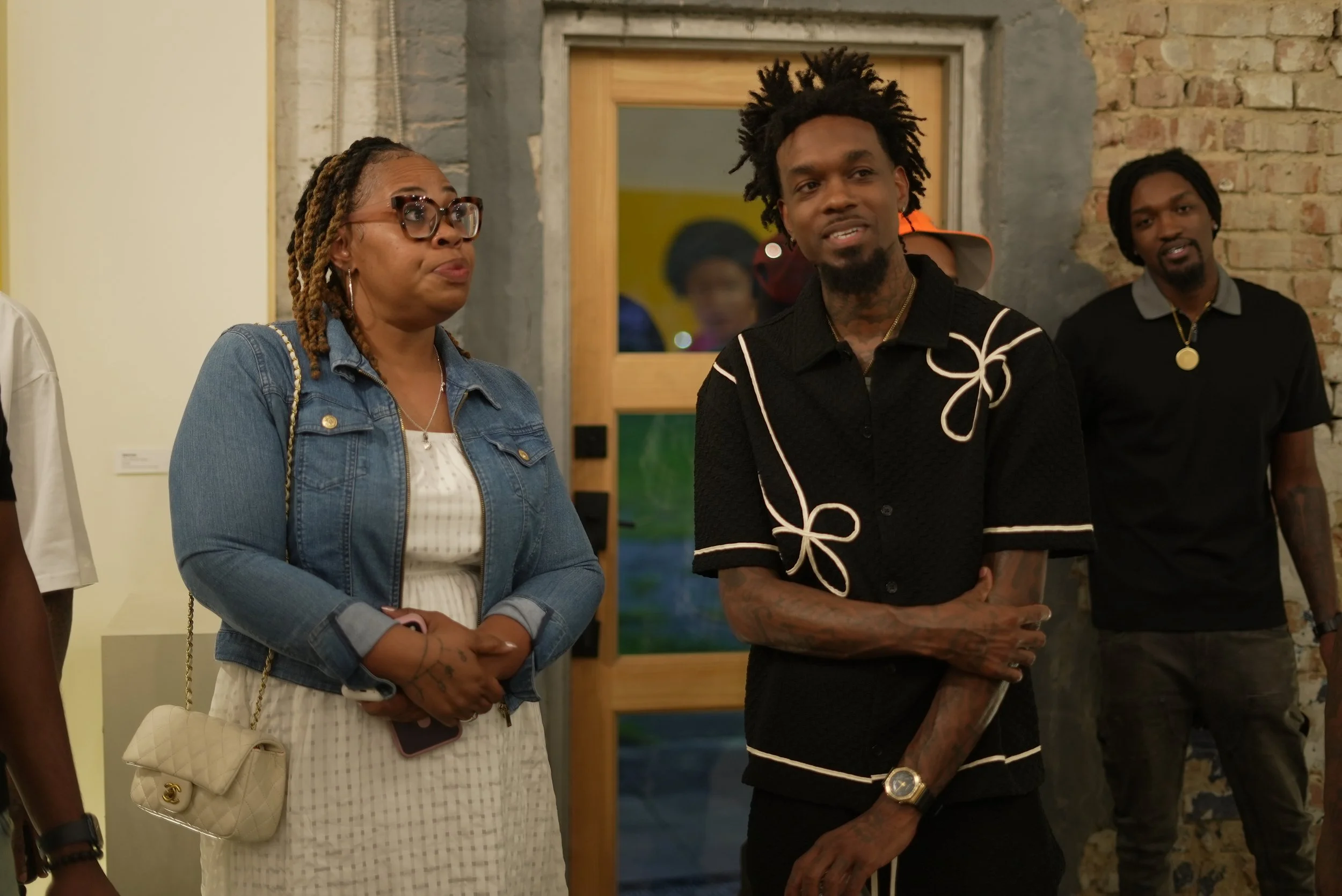 Three people standing indoors at an event, with a brick wall and a wooden door in the background. The woman on the left wears glasses, a denim jacket, and a white dress, holding a phone and a cream-colored purse. The man in the middle has dreadlocks 