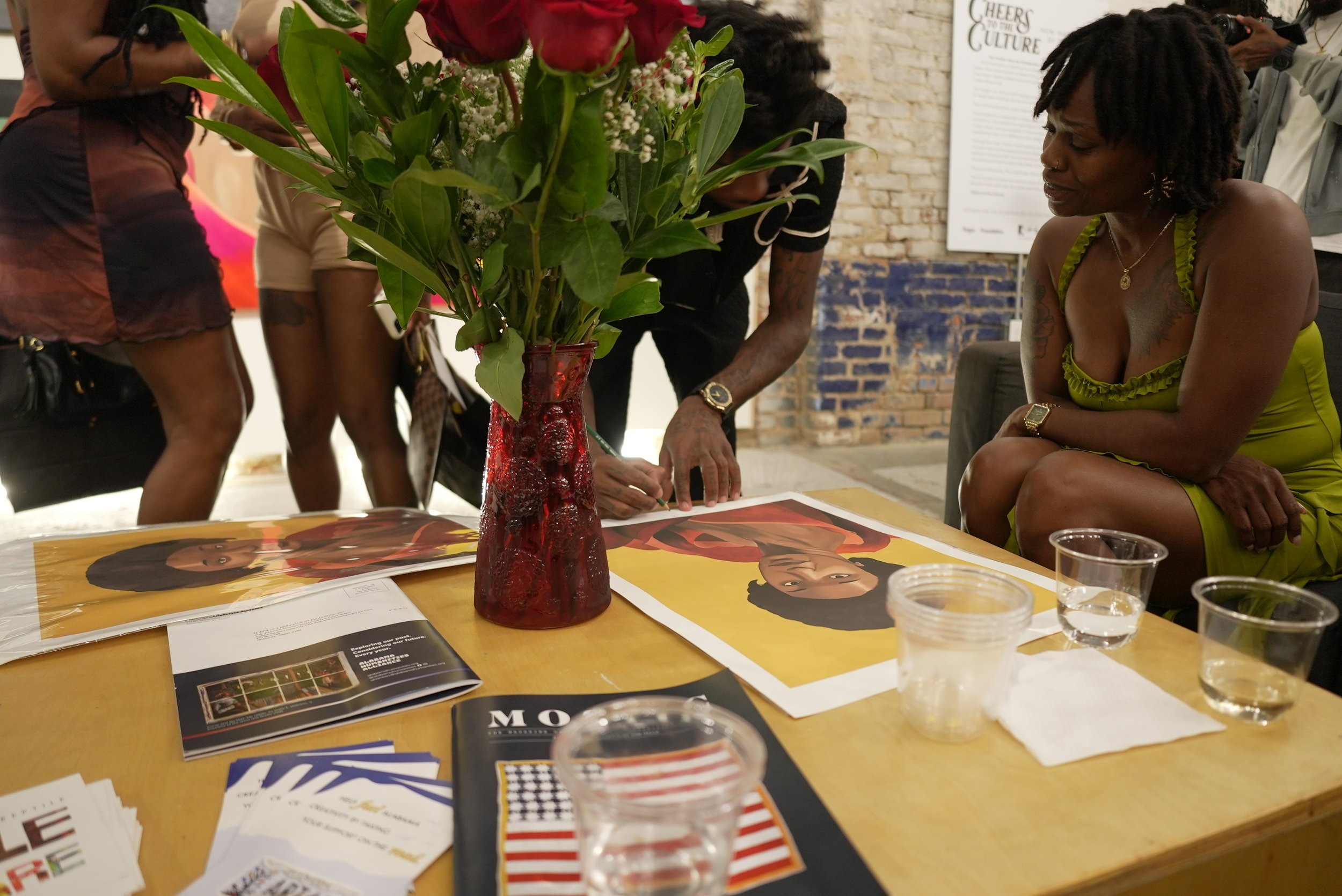 A group of people gathered around a table with an open magazine and various pamphlets, featuring a tall red vase with a large bouquet of green leaves and red flowers, in an indoor setting with a brick wall and framed poster in the background.
