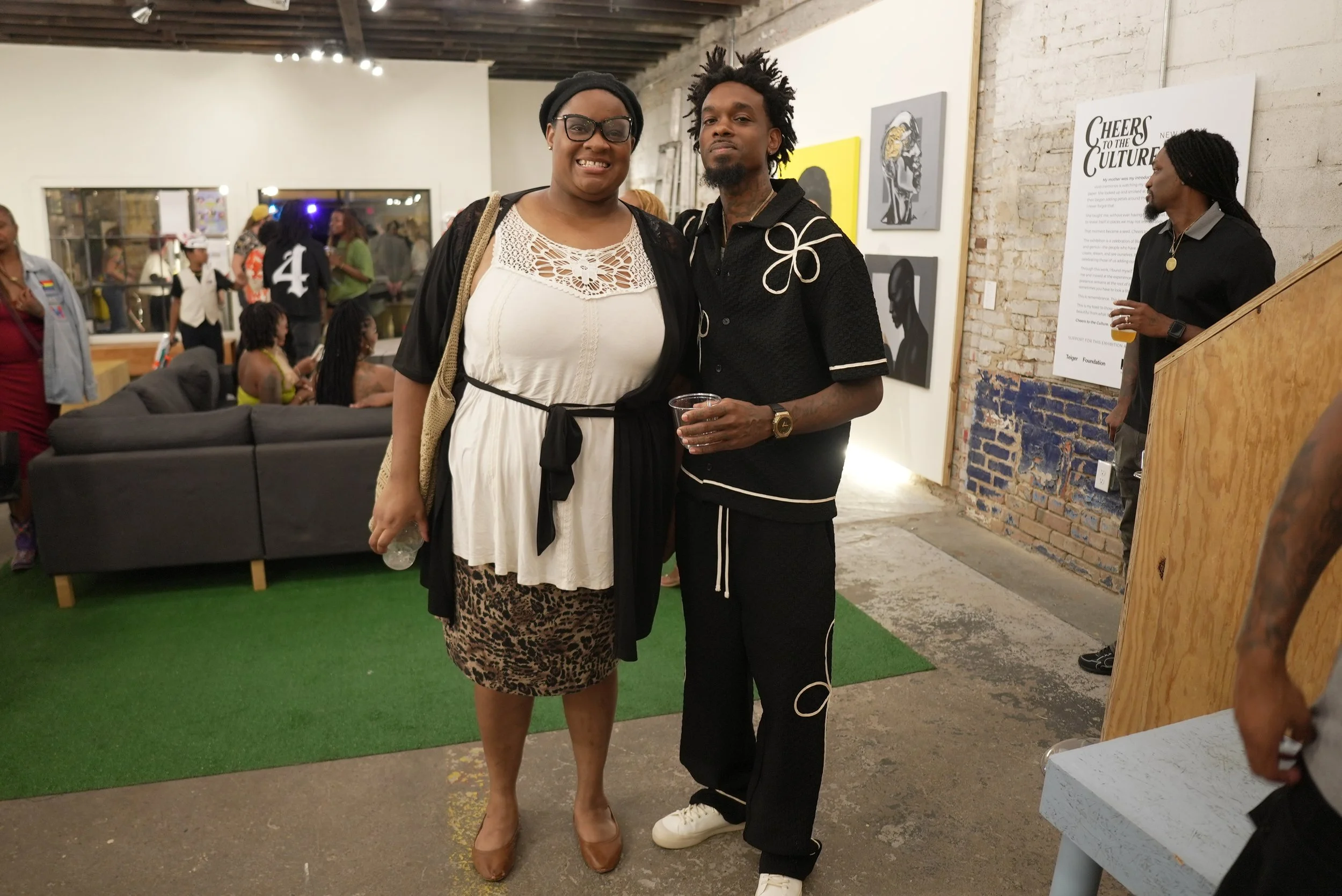 Two women stand together at an art gallery event, smiling at the camera. The woman on the left wears a white dress with lace details, glasses, and a shoulder bag. The woman on the right has dreadlocks, wears a black outfit with white piping, and hold