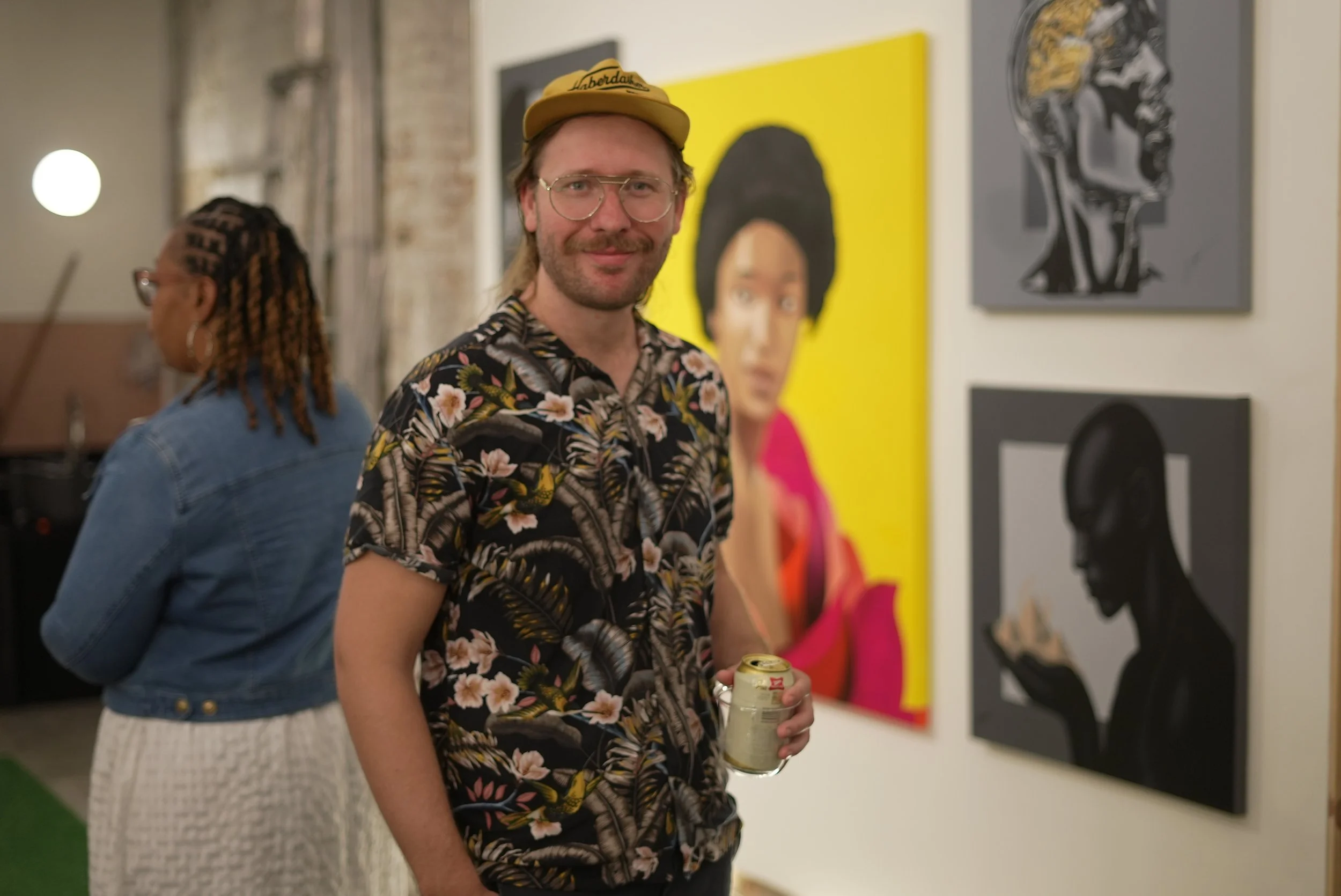 A man with glasses, a beard, and a floral shirt holding a canned drink, standing in an art gallery with colorful portraits on the wall. A woman with braided hair and glasses is in the background.