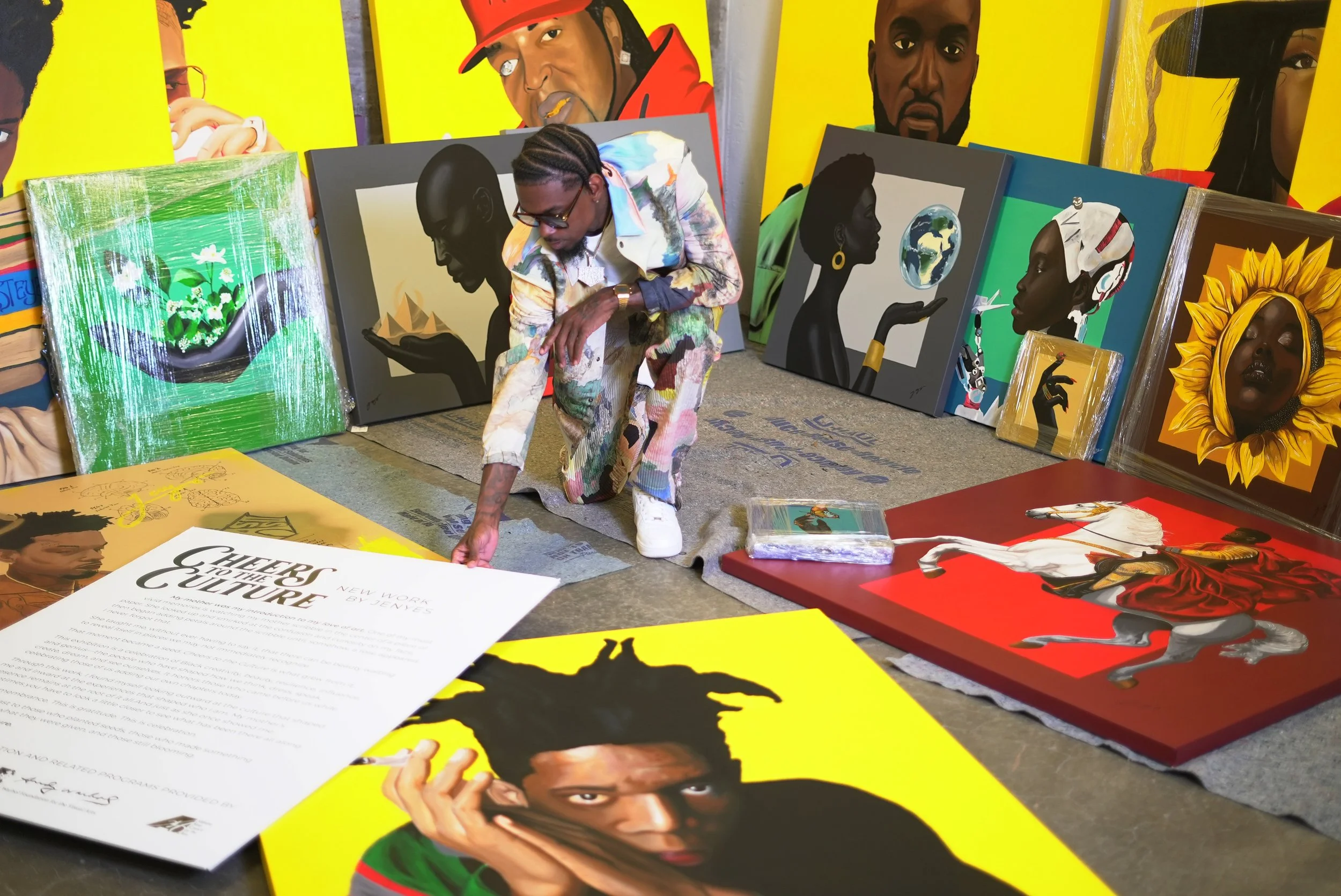 A man in colorful clothing is crouching down to look at art pieces and posters. The background features large, vibrant portraits of people, with some objects like paintings and a printed document on a table in the foreground.