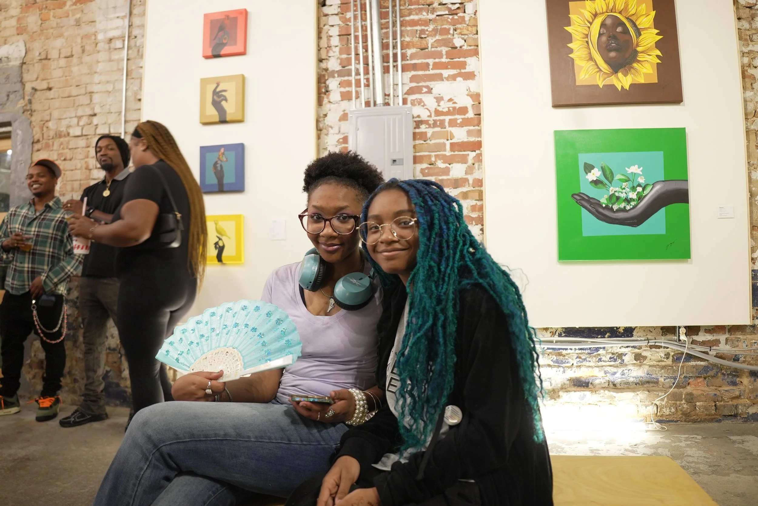 Two women with glasses and colorful hair sitting side by side at an art gallery, smiling at the camera. One holds a handheld fan and wears headphones around her neck. A group of people are standing in the background against brick walls with colorful 