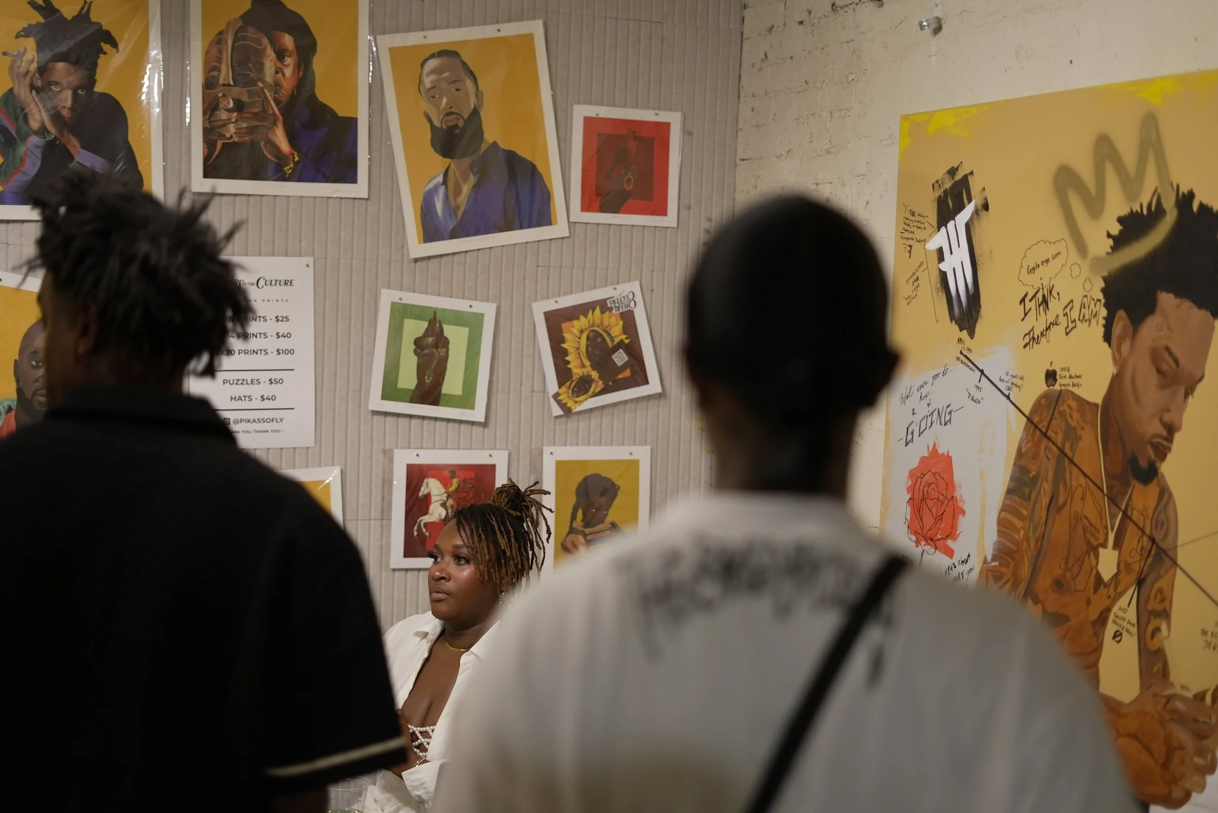 People viewing portrait artwork displayed on a gallery wall, including various images of Black individuals, some with murals or painted styles, with people standing in front of the artwork.