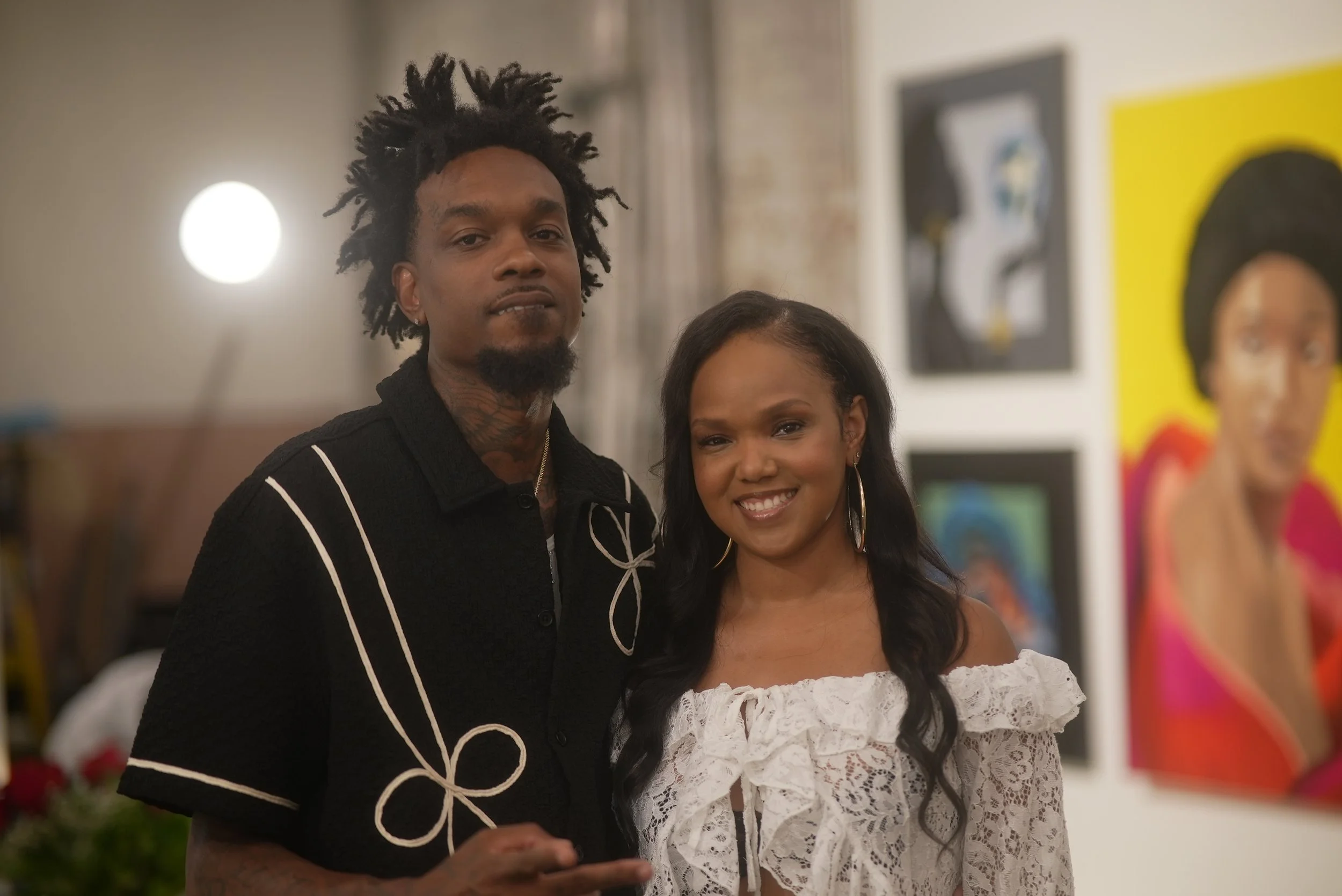 A man and a woman posing together at an art gallery, with artwork displayed on the wall behind them.