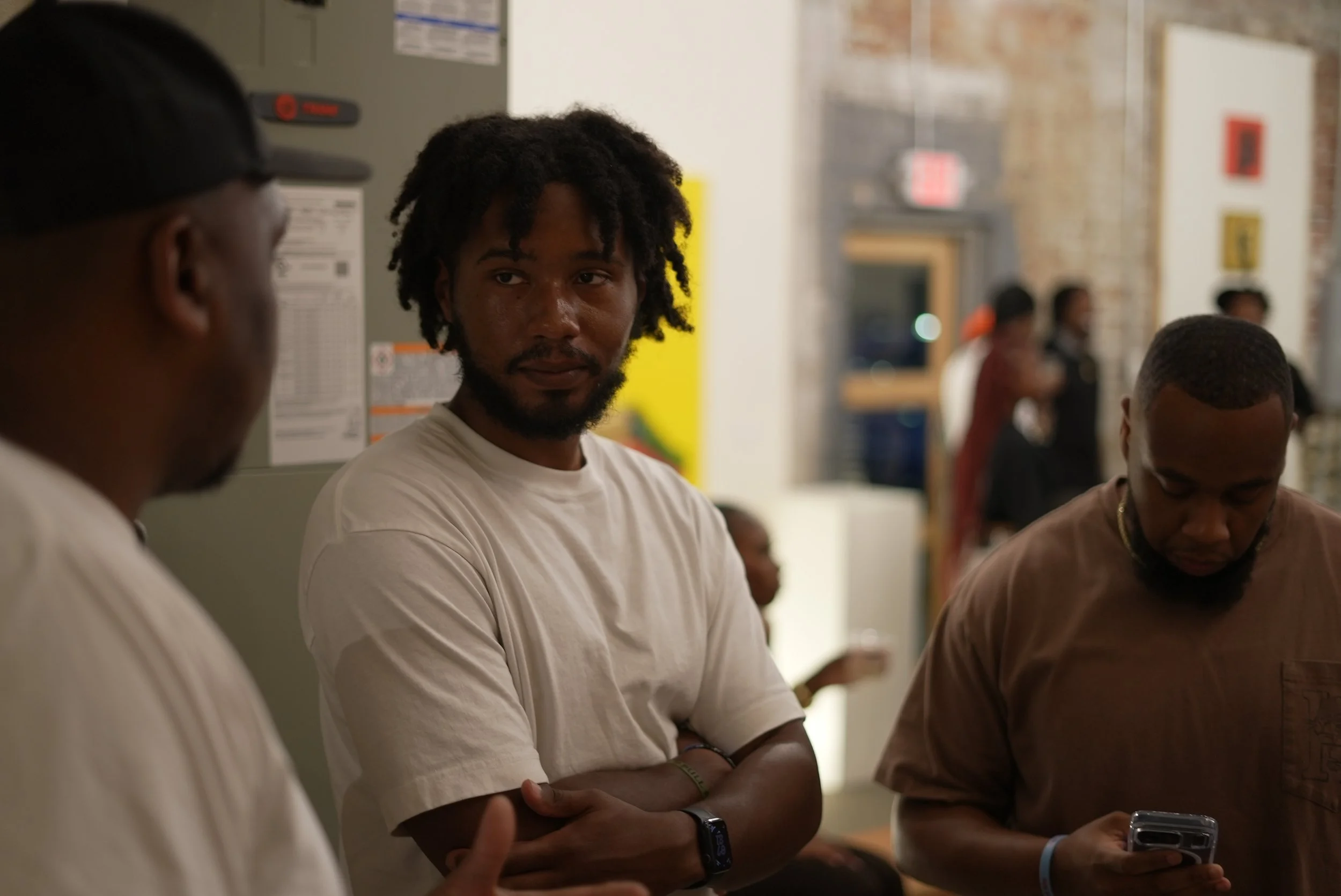 A group of young men are standing indoors, some talking and one looking directly at the camera. The background shows a brick wall and some people in the distance.