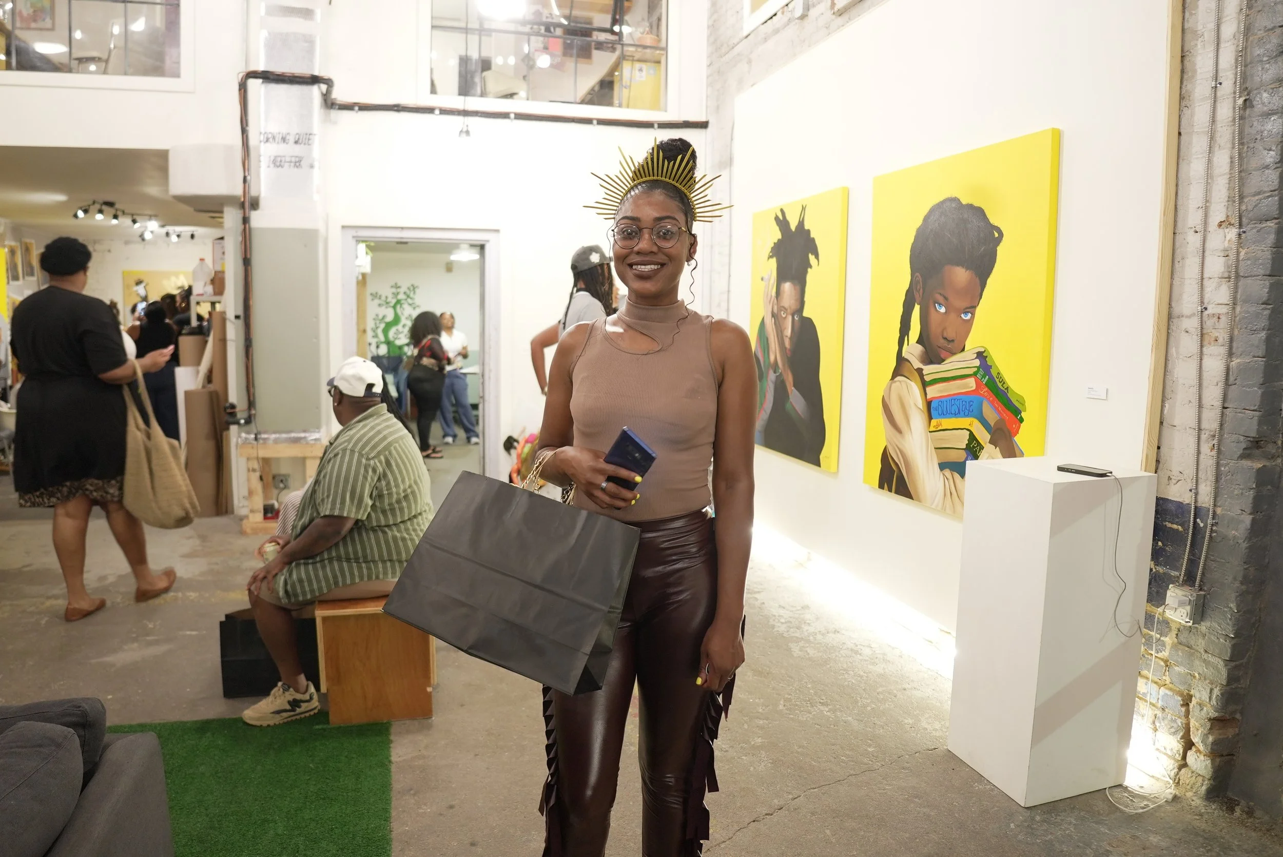A woman with dark skin, glasses, and a golden crown stands inside an art gallery, smiling and holding a black shopping bag. Behind her are colorful portraits of women with diverse hairstyles on yellow canvases. The gallery has white walls, exposed br