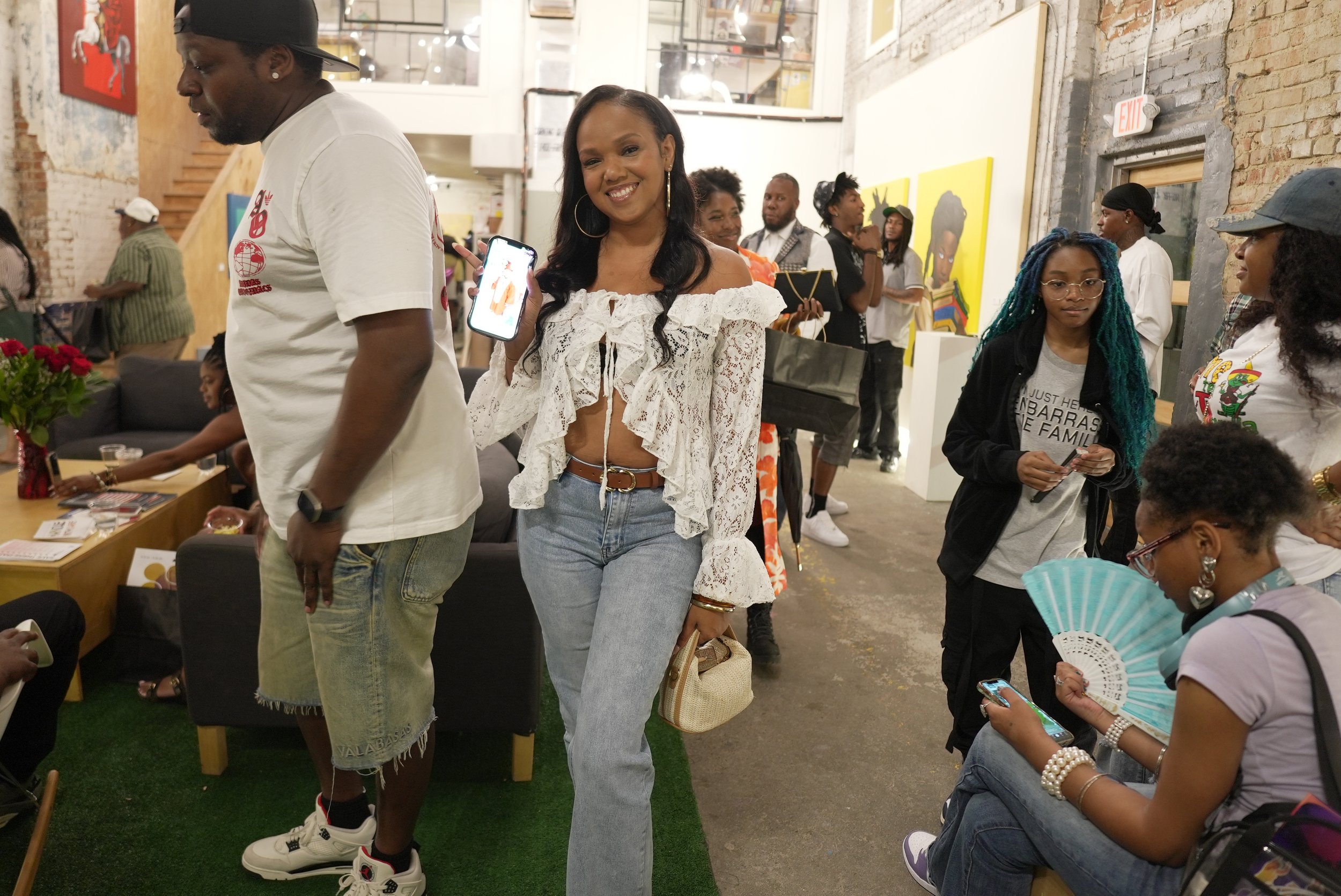 People gathered at an art gallery, woman smiling at camera holding a phone, others looking at artwork, brick walls, colorful paintings