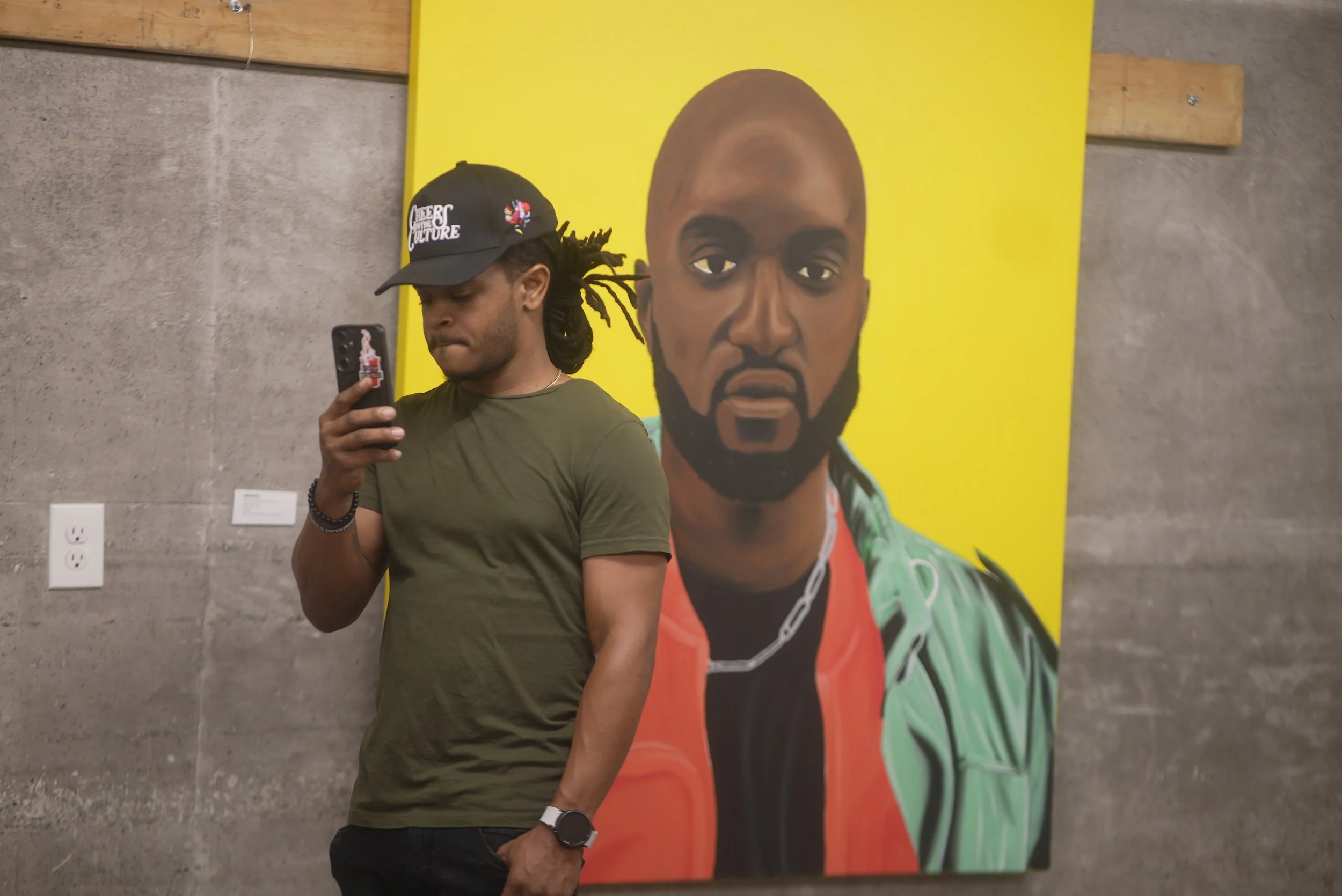 A man with dreadlocks wearing a dark cap and green t-shirt is looking at his phone, standing in front of a large colorful portrait of a bald man with a beard on a yellow background.