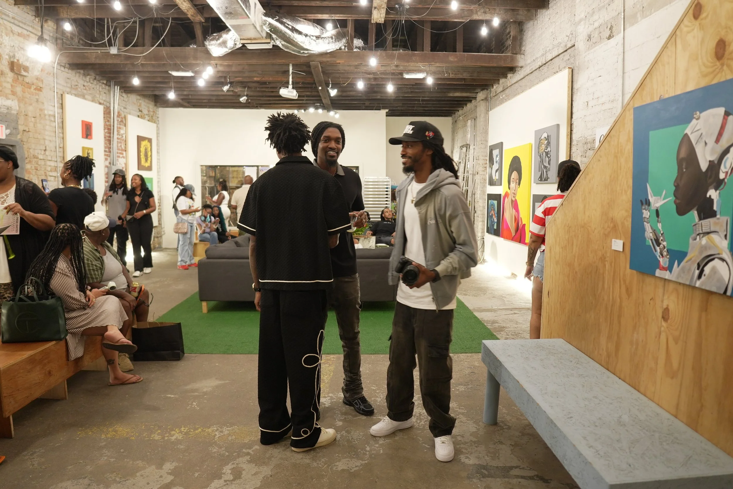 A group of people at an art gallery with paintings on the walls. Three men are in the center talking, one holding a camera. Other visitors are looking at artwork and socializing.